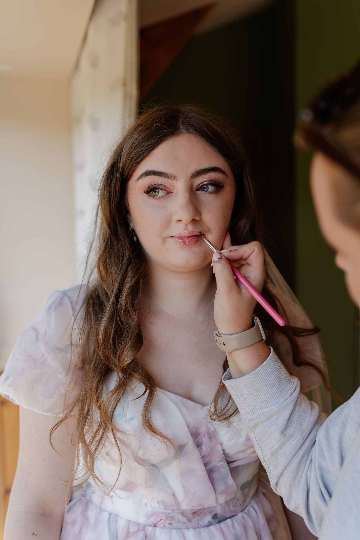 Makeup artist applying lipstick to a young woman with wavy brown hair during bridal preparations.