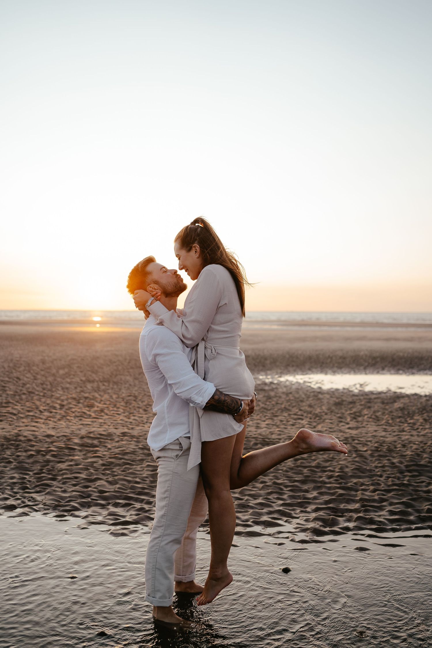 Man lifting woman on a wet beach at sunset, both laughing joyfully in coordinating light-colored outfits.