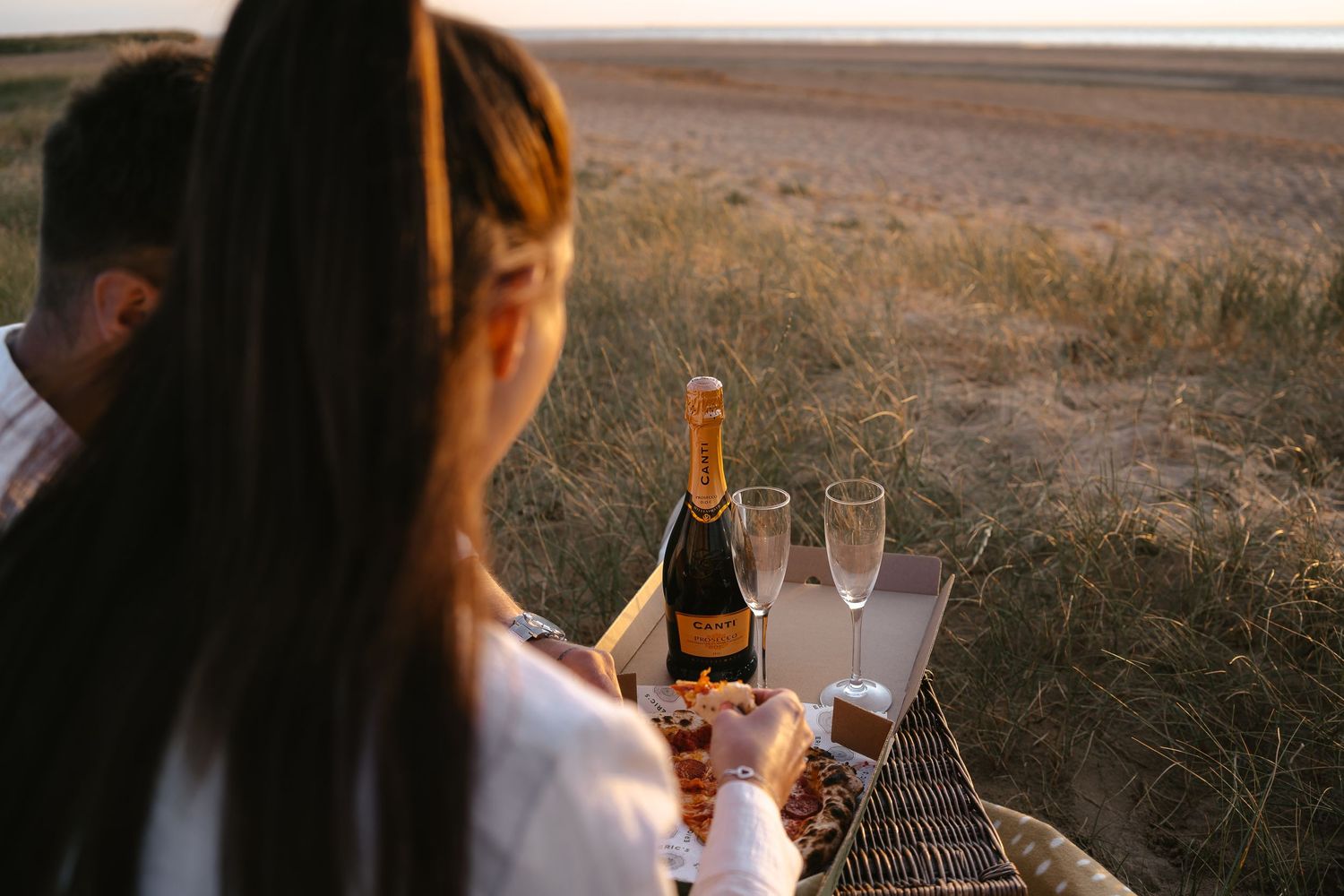 Couple enjoying a romantic beach picnic at sunset with a bottle of Canti champagne and flutes.