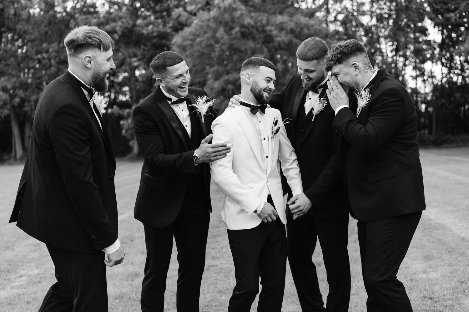 Groom in white tuxedo laughs with groomsmen in black suits in a black and white outdoor photo.