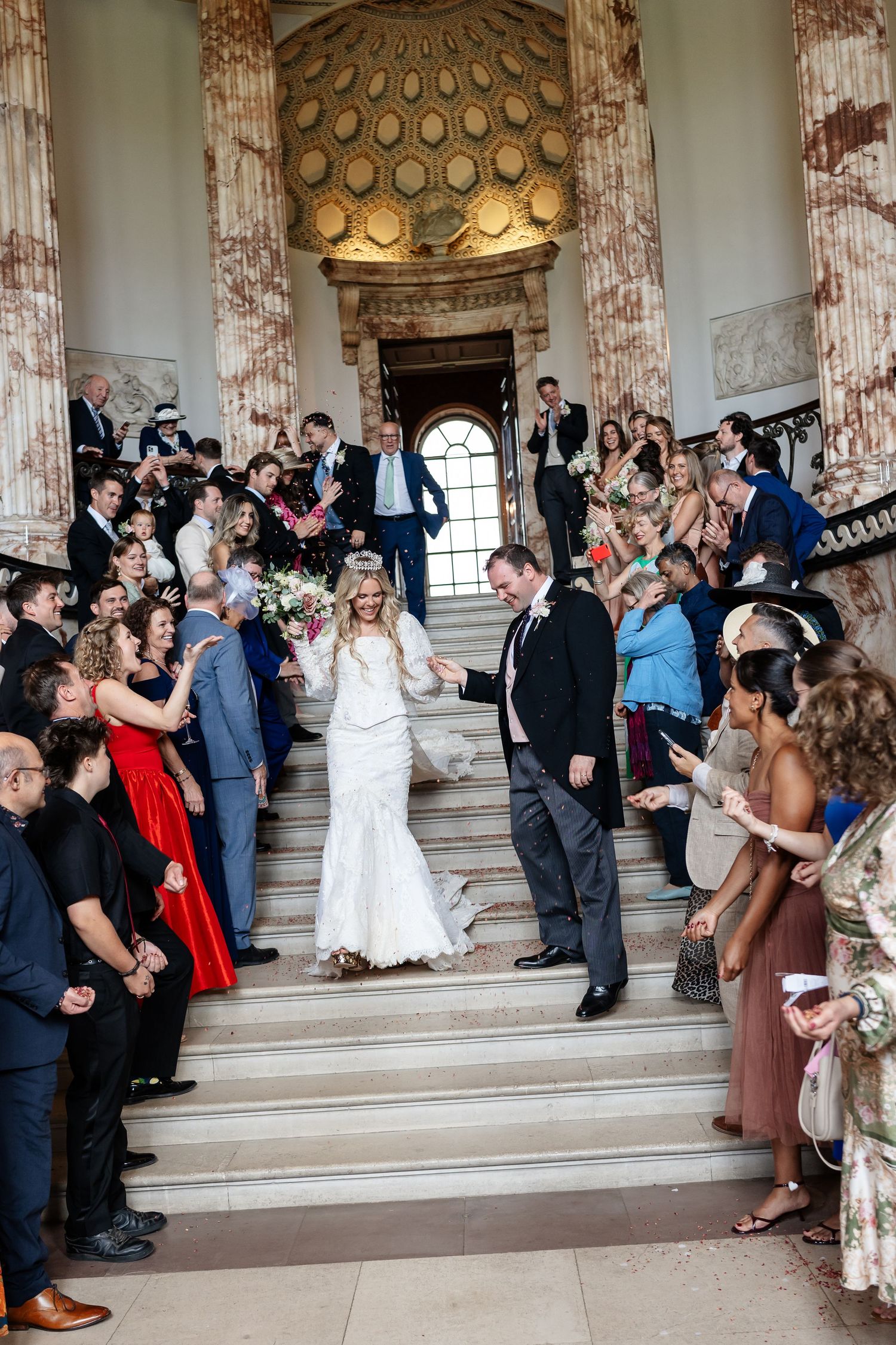 Newlyweds descend grand staircase surrounded by cheering wedding guests in stunning neoclassical venue.