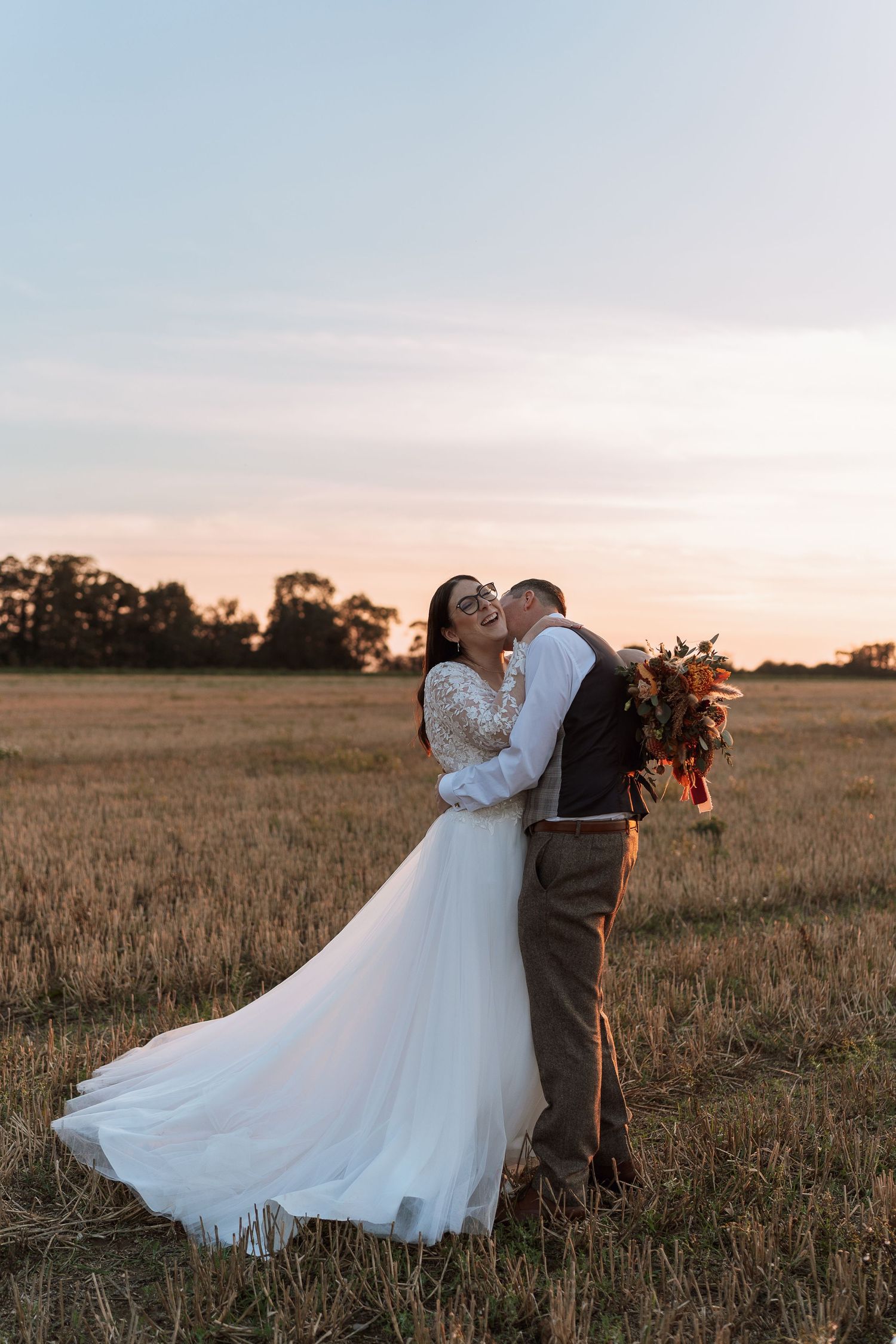 A bride and groom embrace romantically in a golden wheat field at sunset on their wedding day.