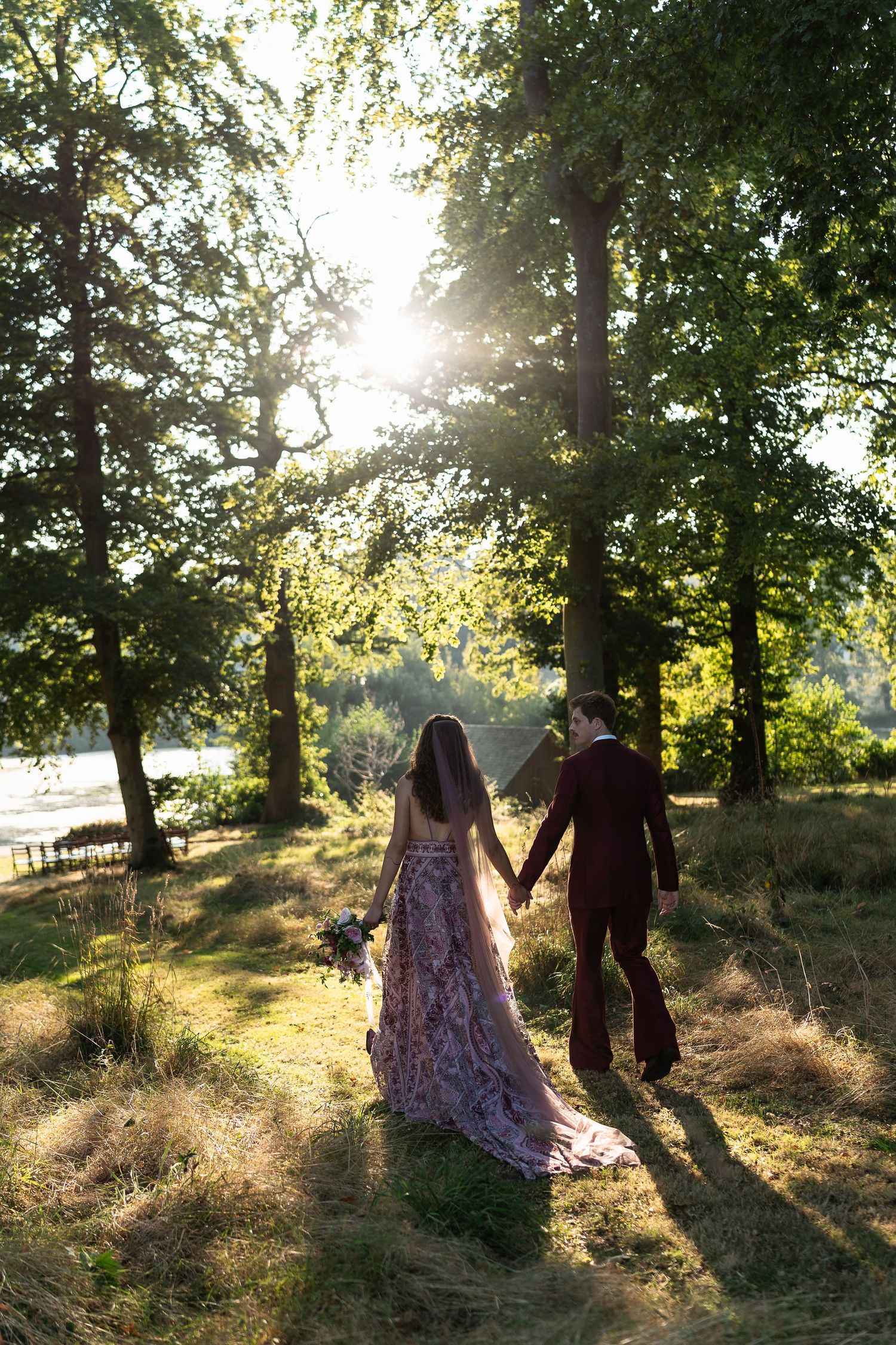 Bride in pink floral gown and groom in burgundy suit walk hand in hand through sunlit woodland.