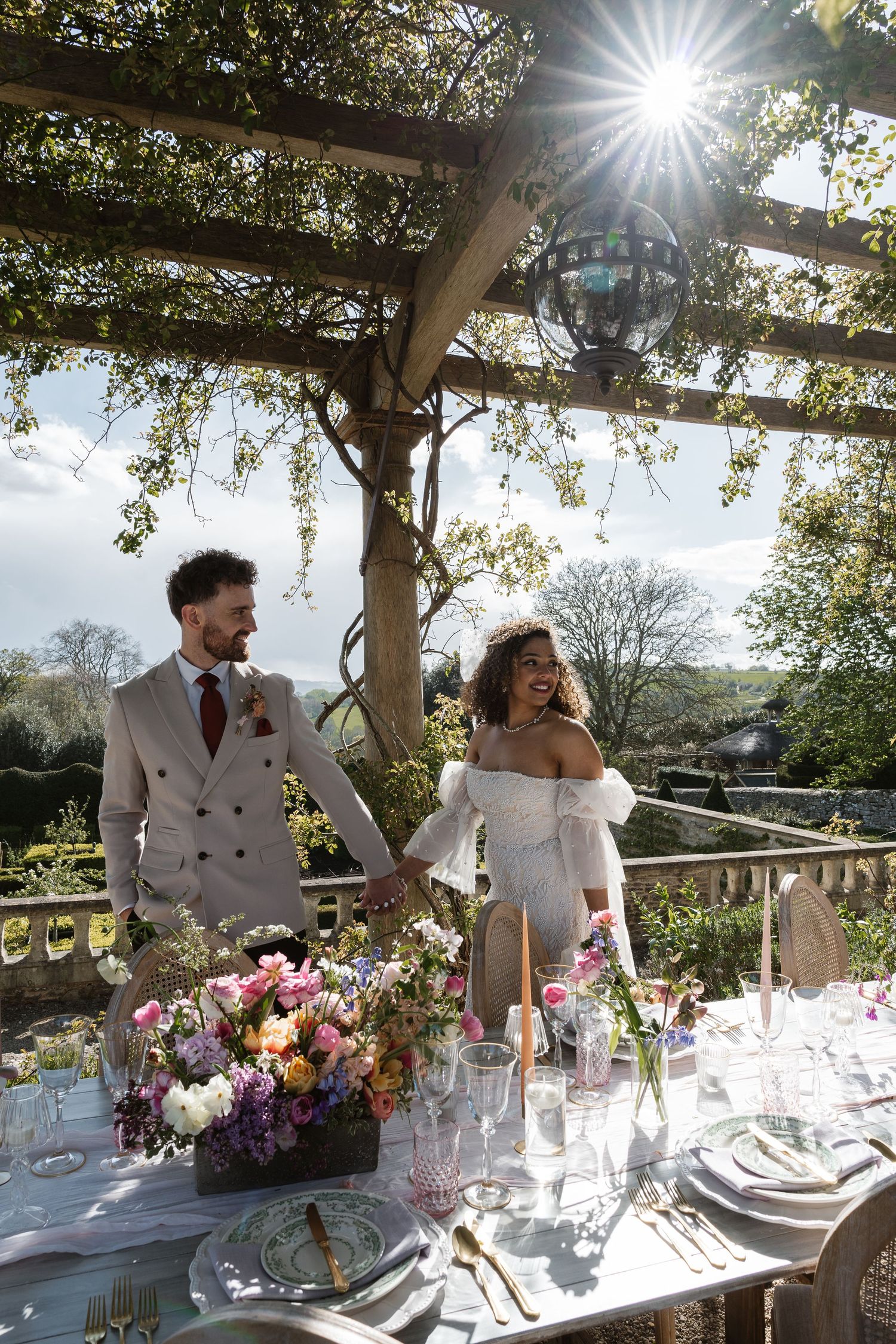 Couple holding hands at outdoor wedding reception table with colorful floral centerpiece under pergola in sunlight.