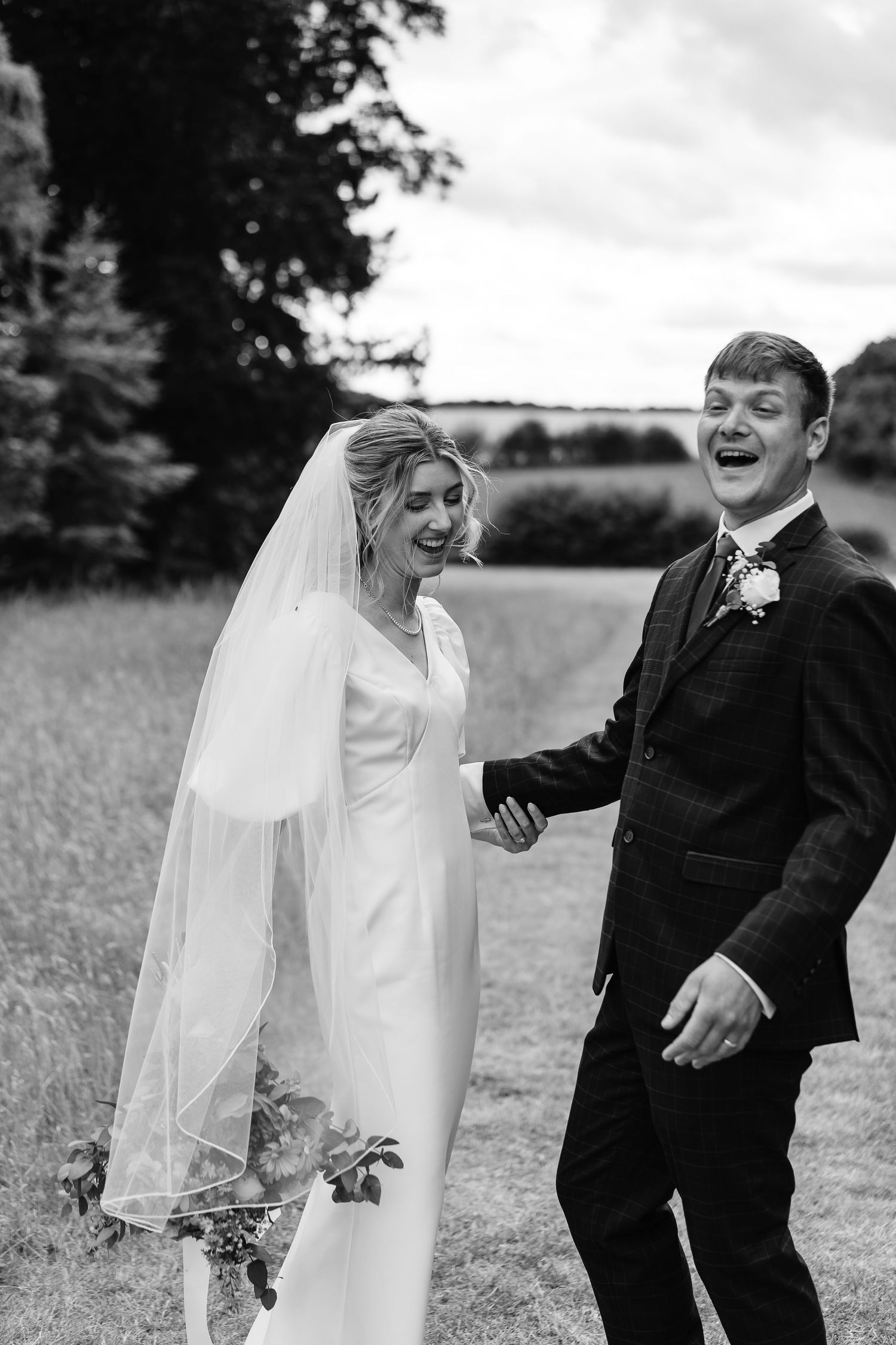 Laughing bride and groom walk hand in hand through countryside field in black and white photo.