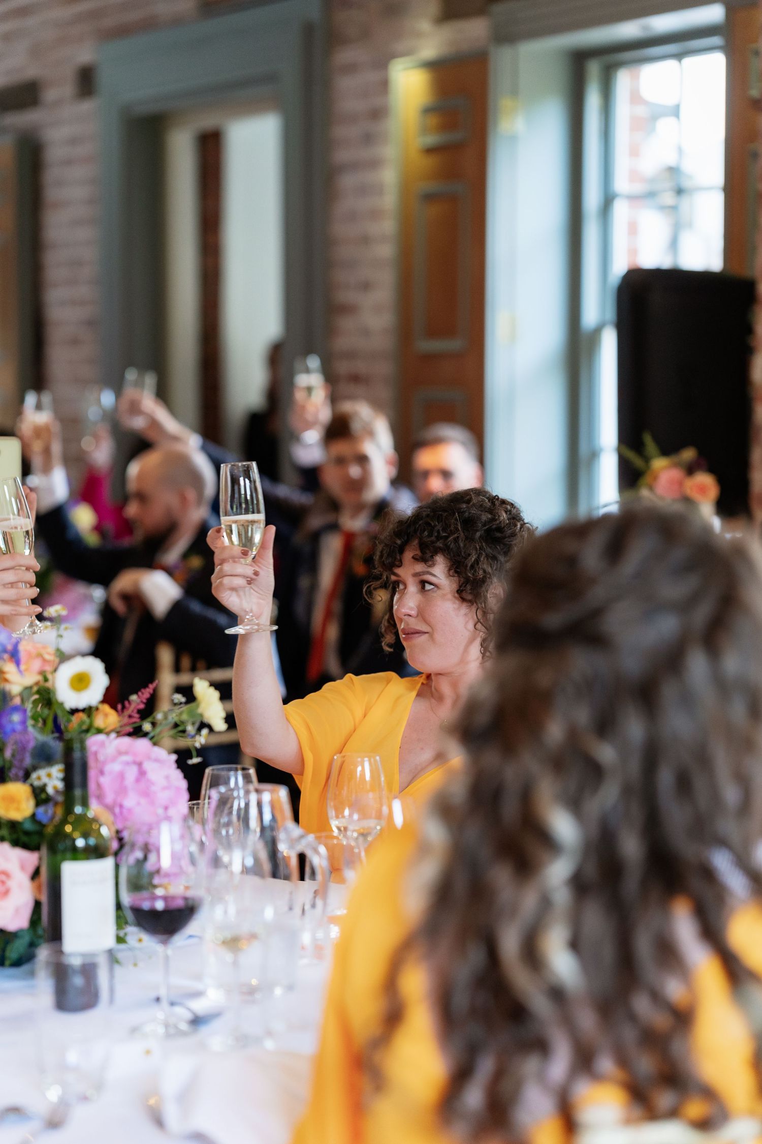 Wedding guest in yellow dress raises champagne glass during a toast at a floral-decorated reception table.