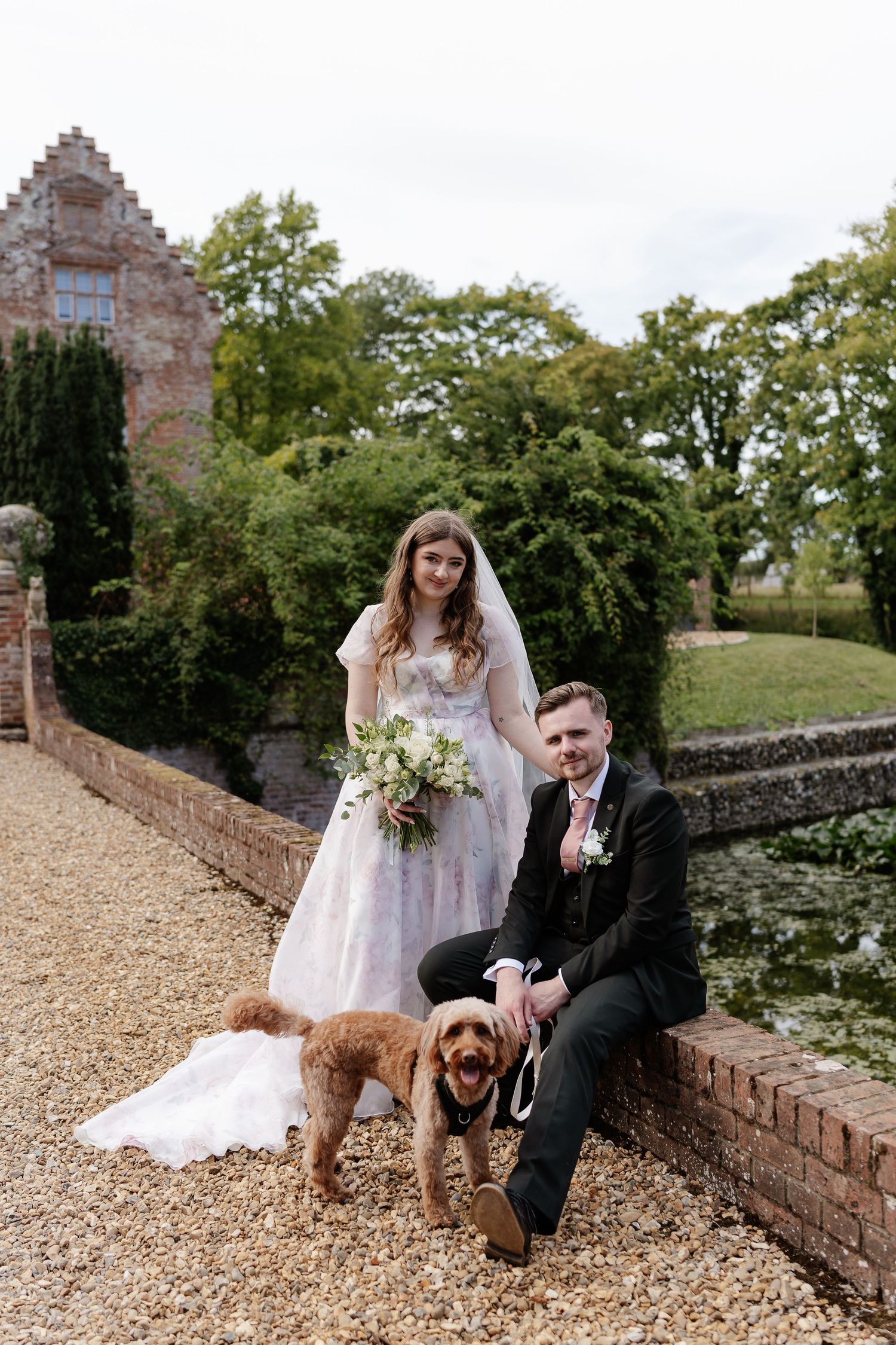 Bride and groom pose with golden doodle dog beside brick wall and pond at elegant wedding venue.