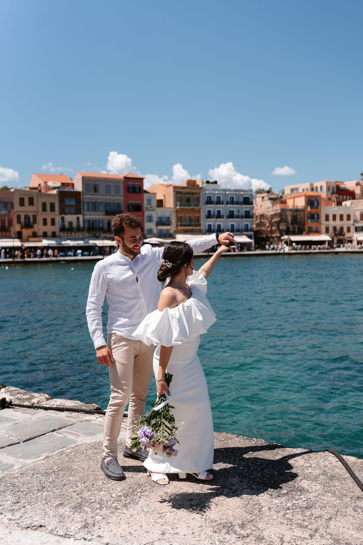 Bride in white off-shoulder dress spins on a Mediterranean harbor dock as groom watches, colorful buildings in background.