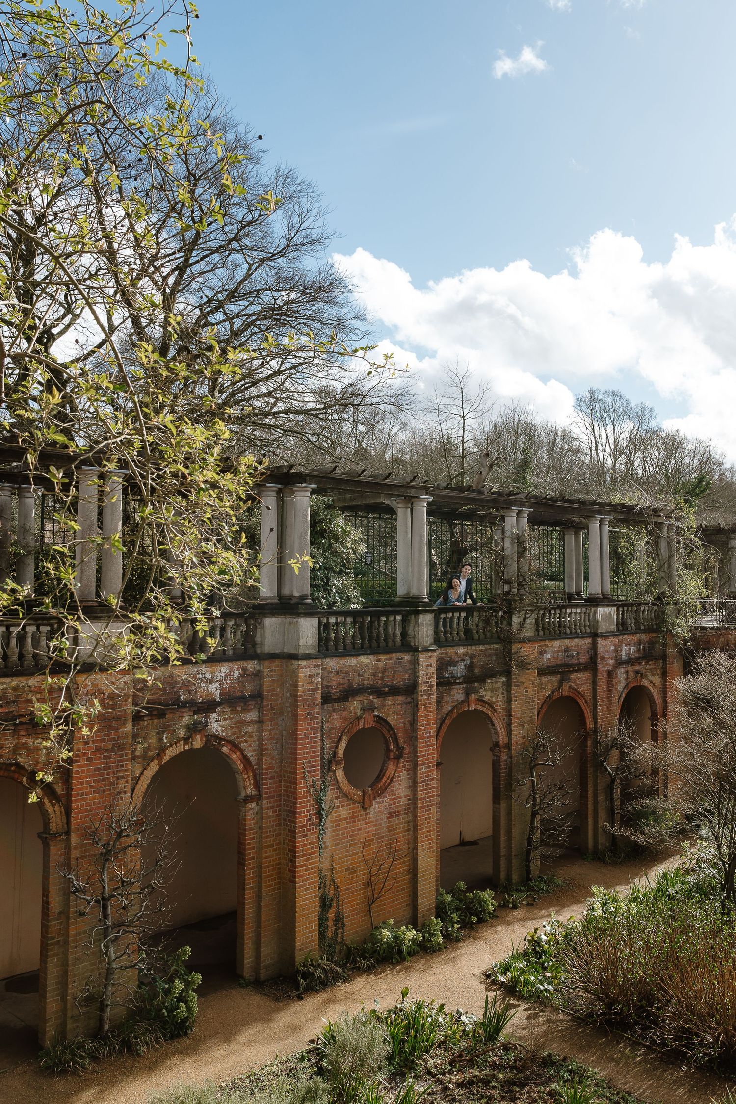 Historic brick arcade with arched openings and columned terrace surrounded by trees under a cloudy sky.