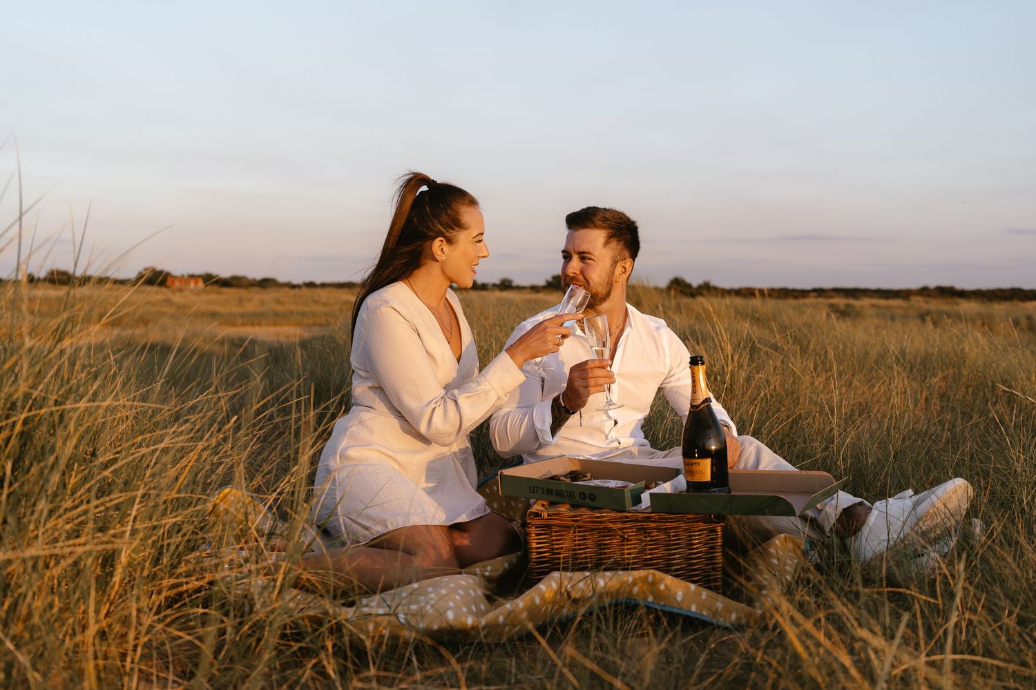 Romantic couple enjoying a picnic with champagne and pizza in a golden wheat field at sunset.