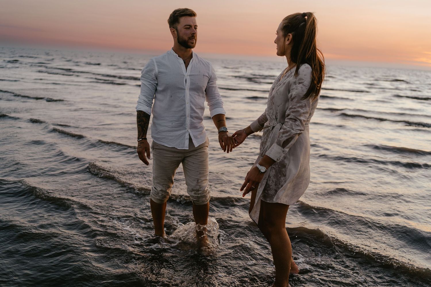 Couple holding hands walking in ocean waves at sunset, wearing white outfits on a romantic beach.
