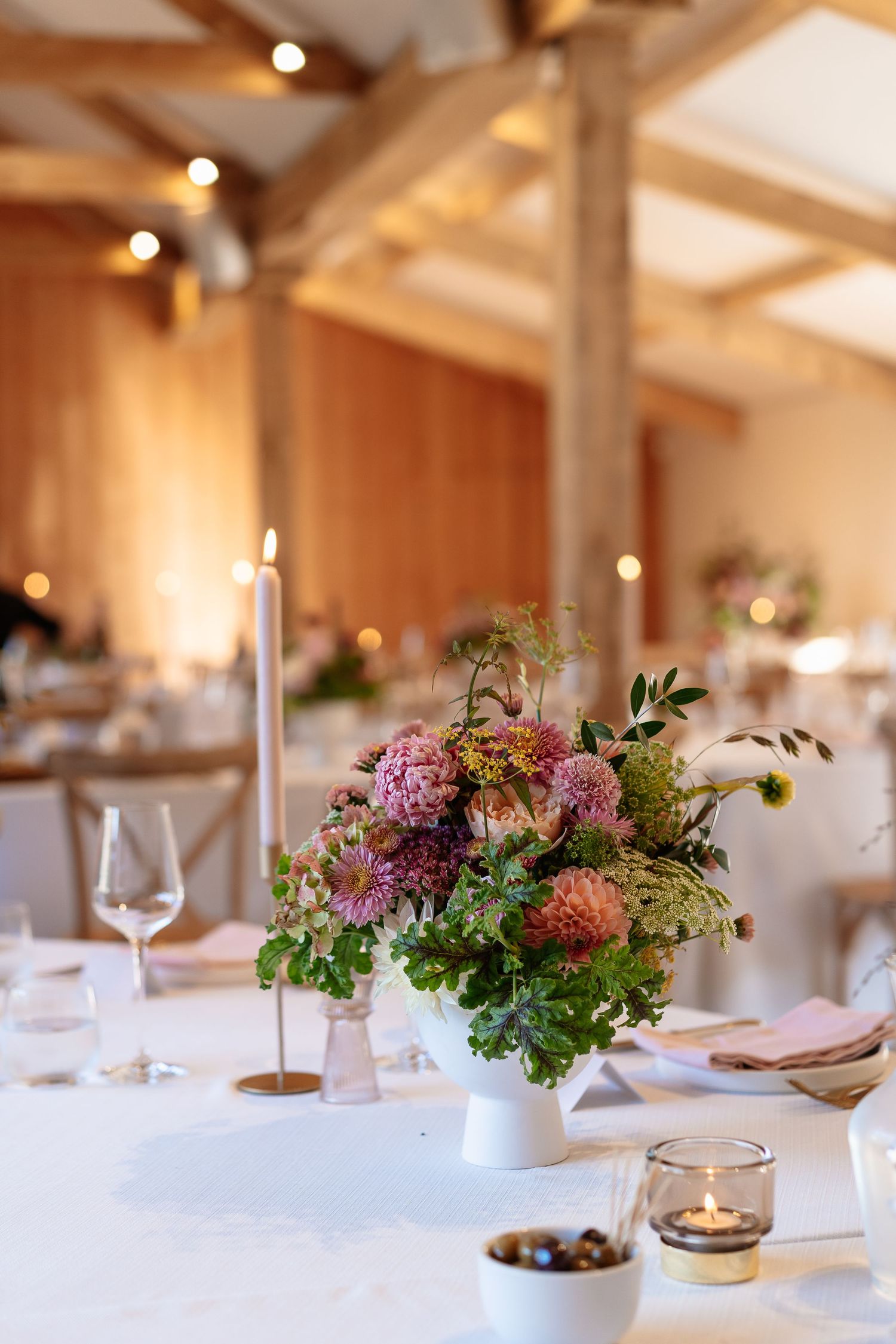 Elegant wedding reception table with floral centerpiece, candles, and glassware in a warm barn venue.