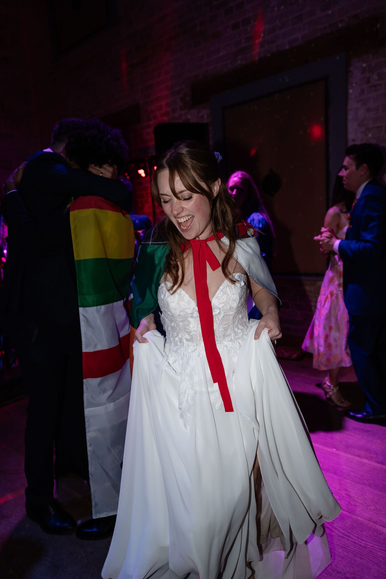 A joyful bride in a white gown with red ribbon dances at her wedding reception.