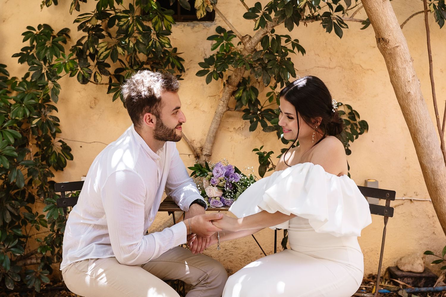 Bride and groom holding hands and smiling at each other in a charming sunlit outdoor courtyard.