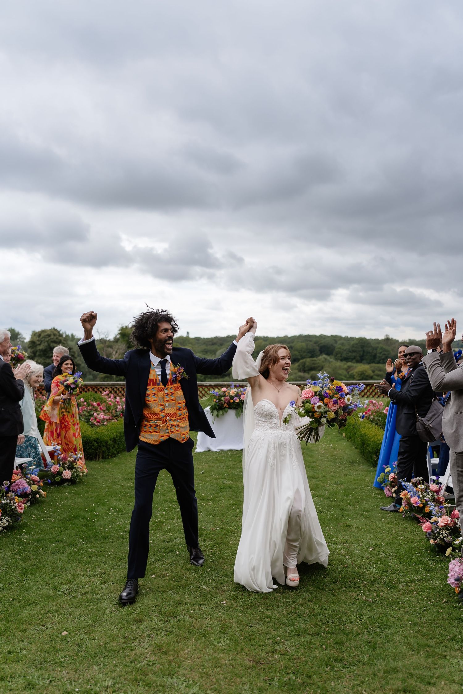 Ecstatic groom jumps with joy as couple raises hands celebrating after outdoor garden wedding ceremony.