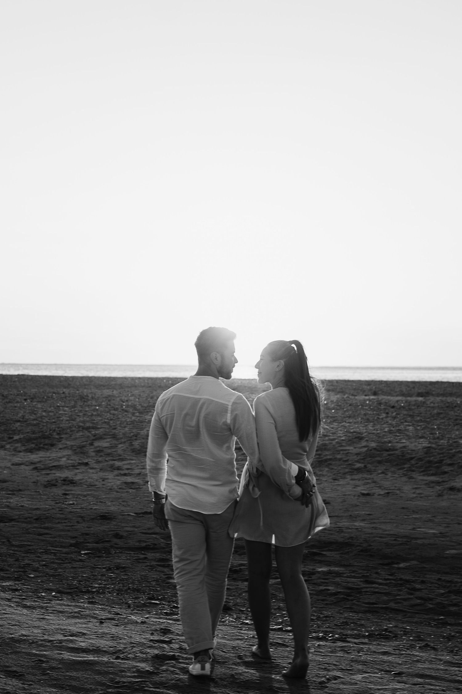 Black and white photo of a couple walking arm in arm along a beach at sunset.