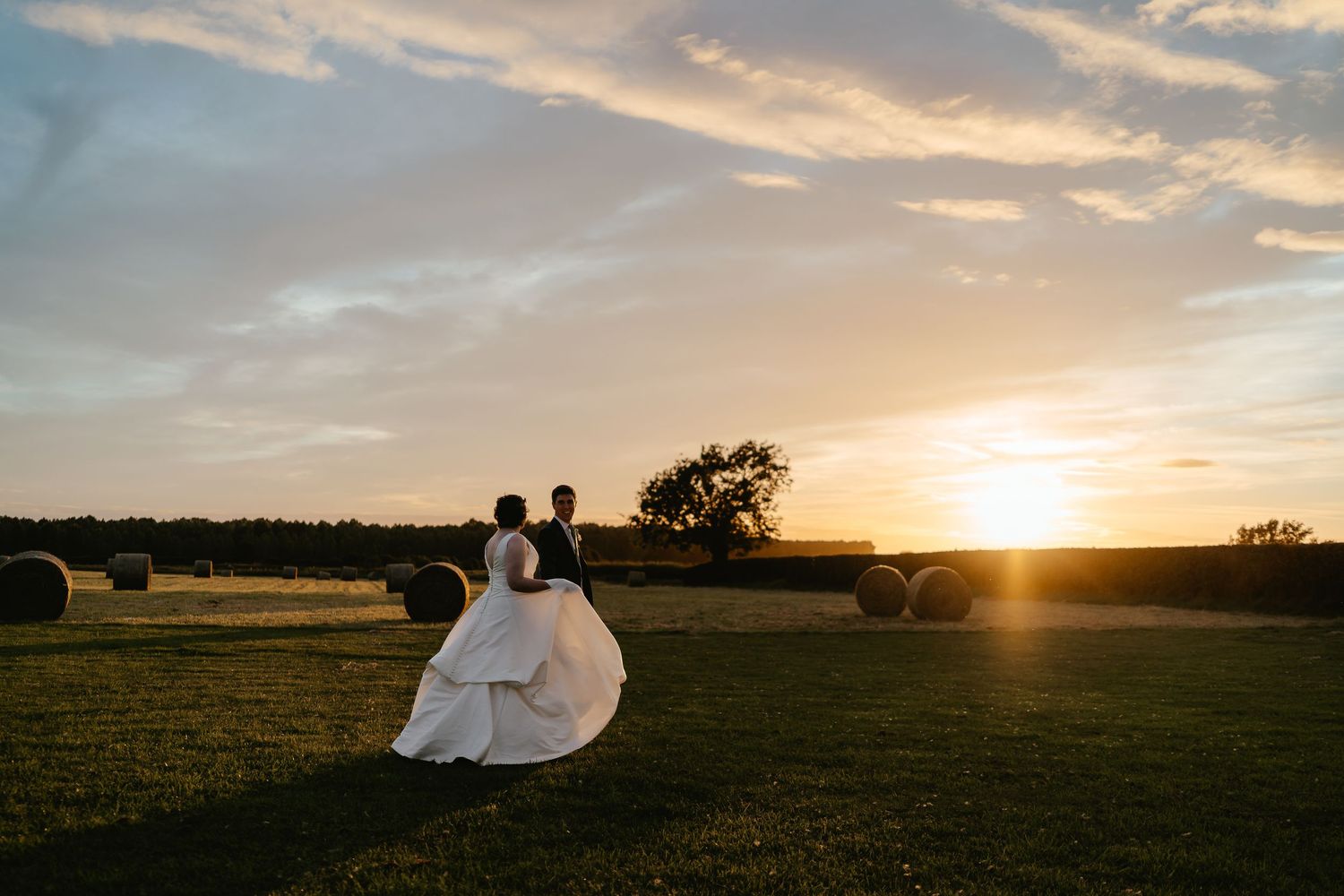 Bride and groom walking during golden hour at Pentney Abbey across the fields with haybales