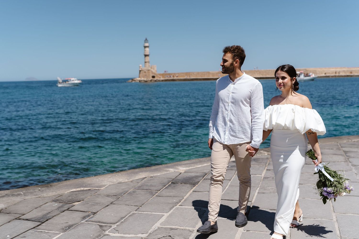 Bride and groom walk hand in hand along a waterfront promenade with a lighthouse and blue sea behind them.