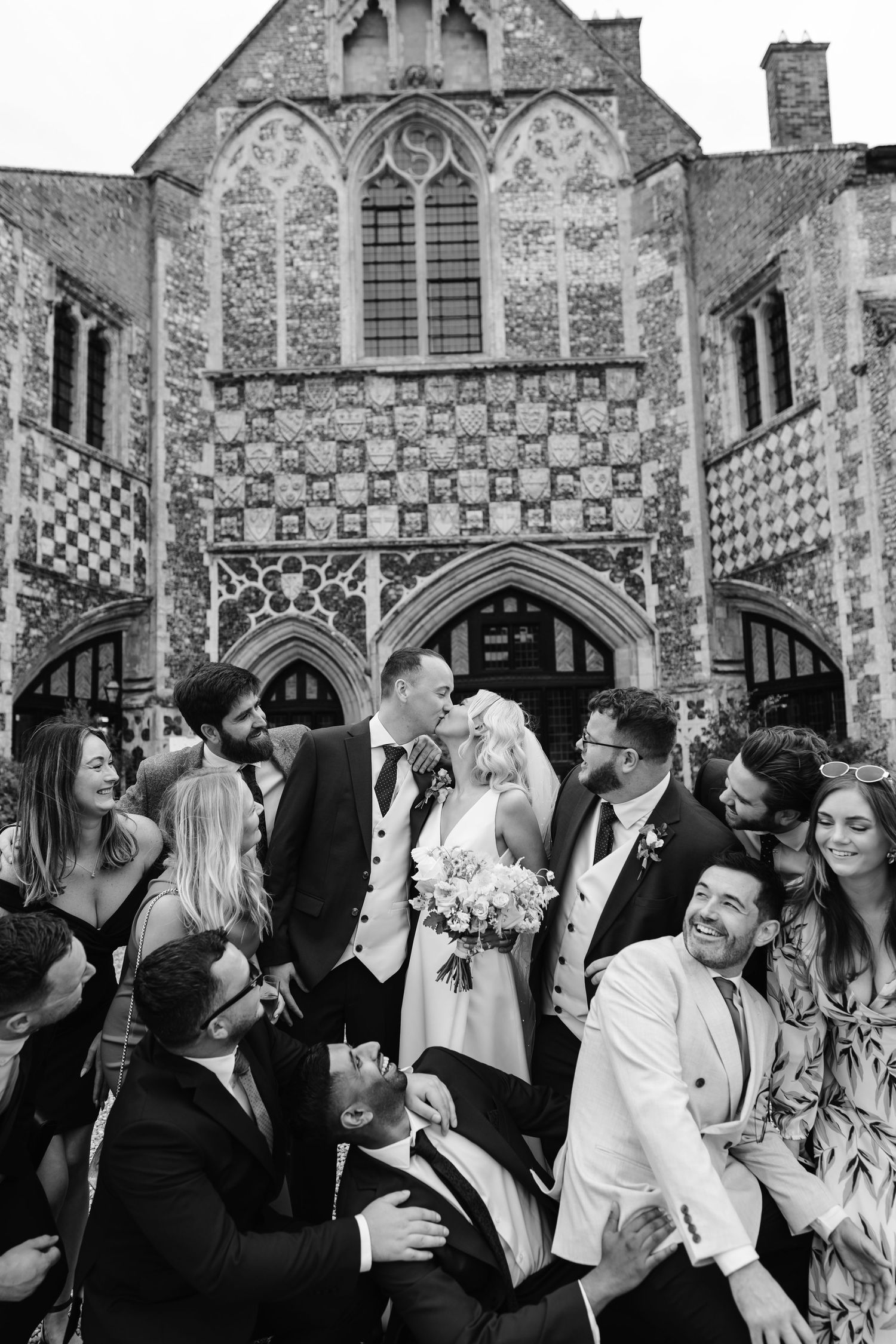 Black and white wedding photo of bride and groom kissing surrounded by guests outside a Gothic stone building.