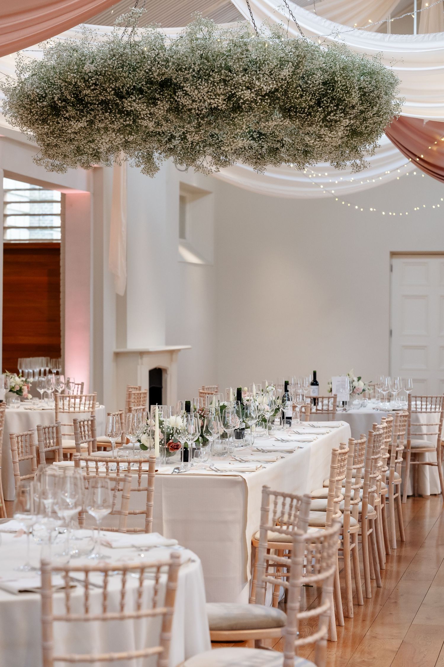 Elegant wedding reception hall with baby's breath floral chandelier, white tables, chiavari chairs, and fairy lights.