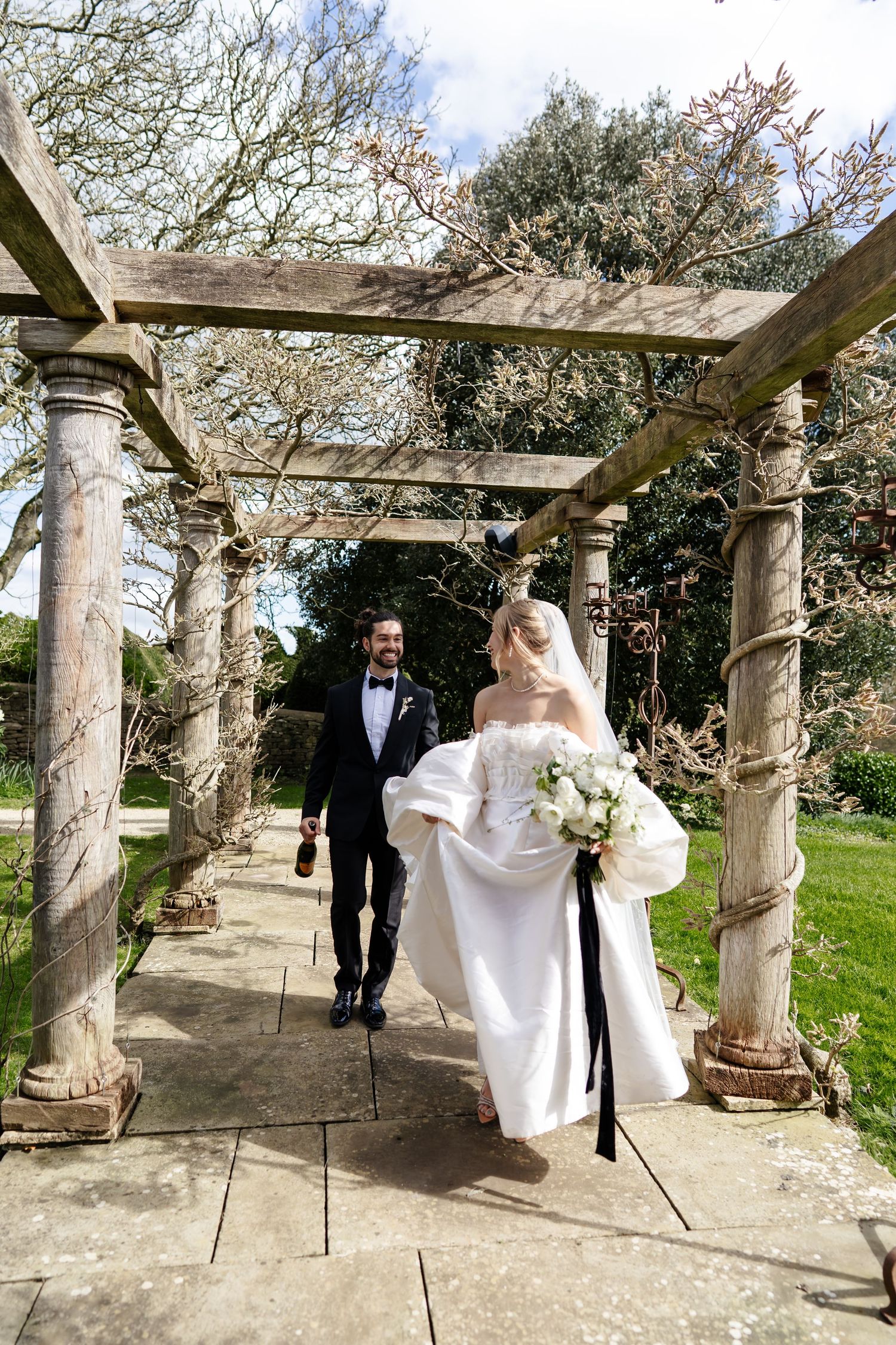 Bride and groom walking hand in hand under rustic wooden pergola in spring garden.