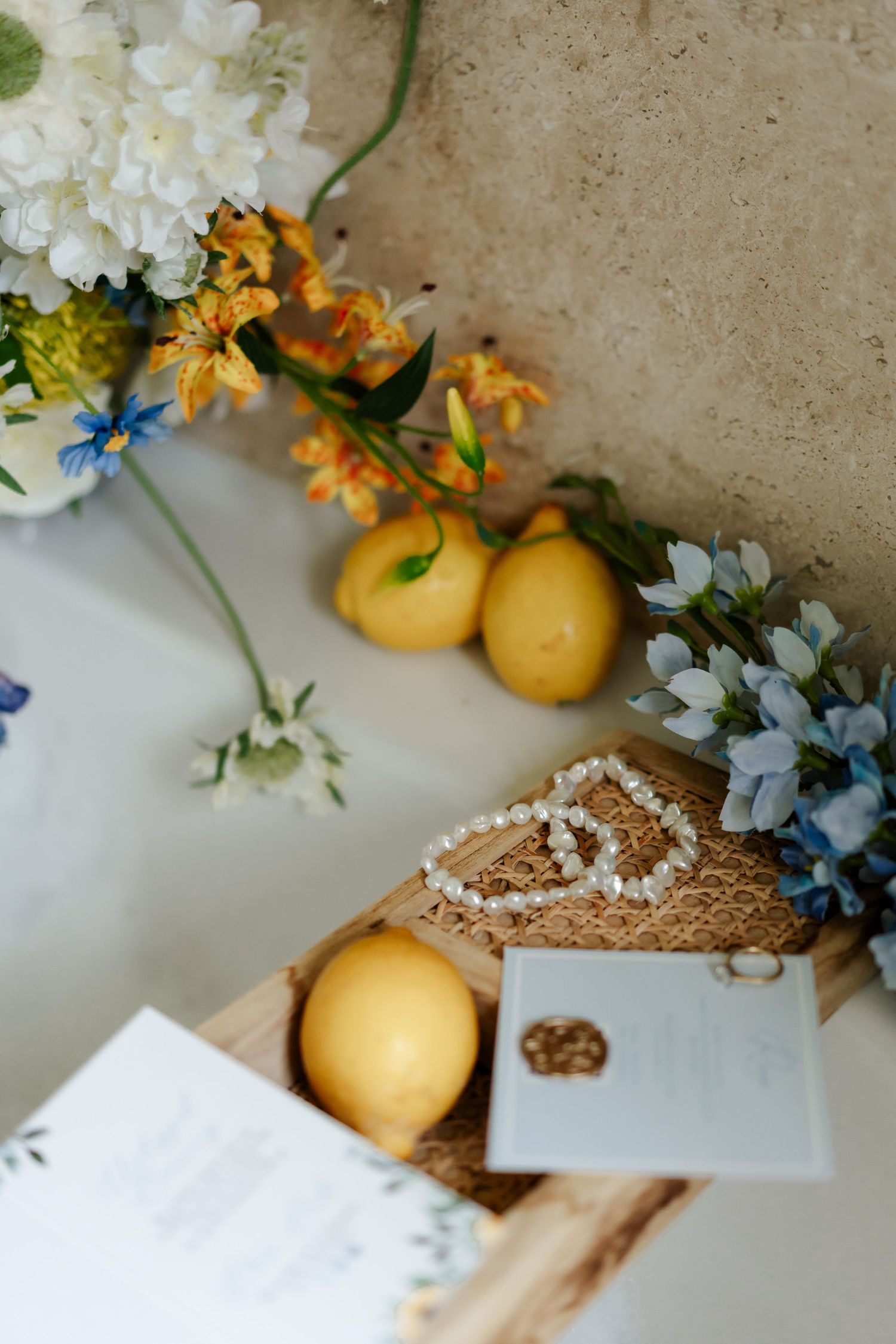 Wedding flat lay with lemons, pearl bracelet, rings, blue flowers, and invitation stationery.