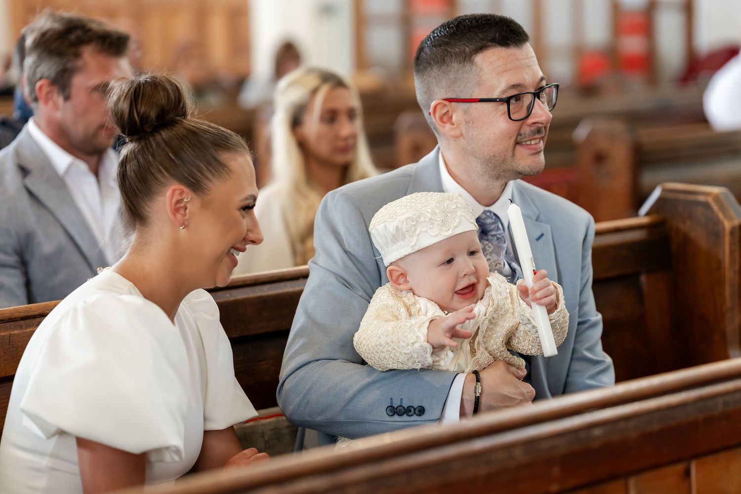 Man in grey suit holds smiling baby at a church christening ceremony beside a woman in white.