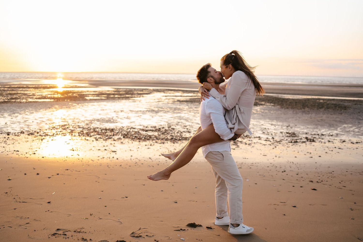Couple sharing romantic kiss as man lifts woman on beach at golden sunset.