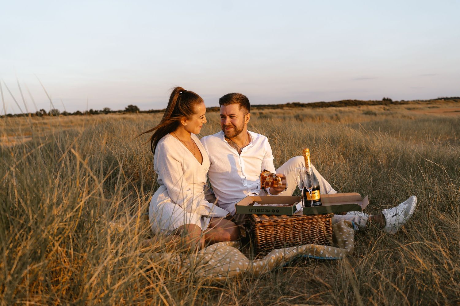 Couple sharing a romantic picnic moment in a vast golden field at sunset.