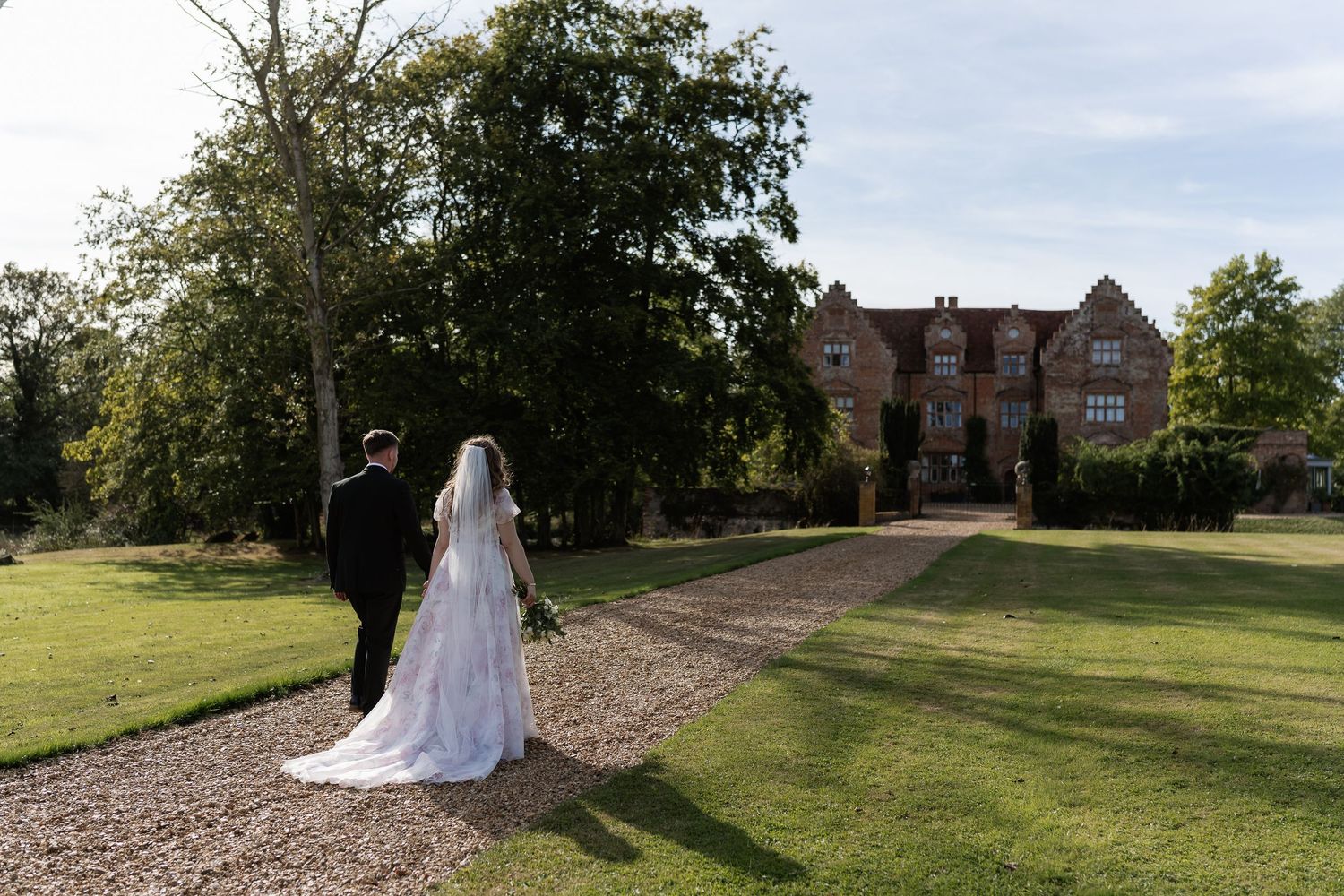 Bride and groom walking hand in hand toward a grand red brick manor house on a sunny day.