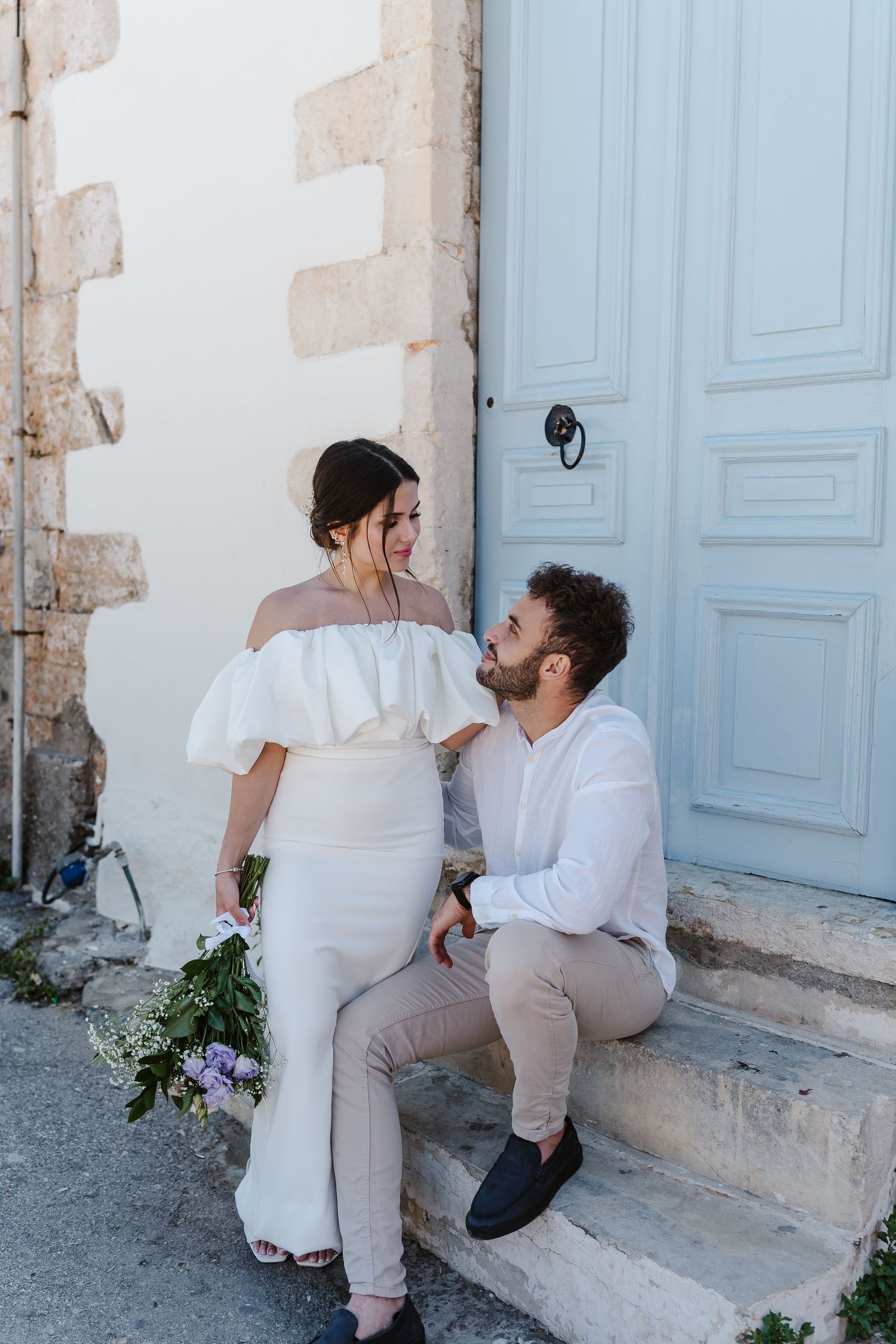 Bride and groom share a loving glance while seated on stone steps by a blue door.
