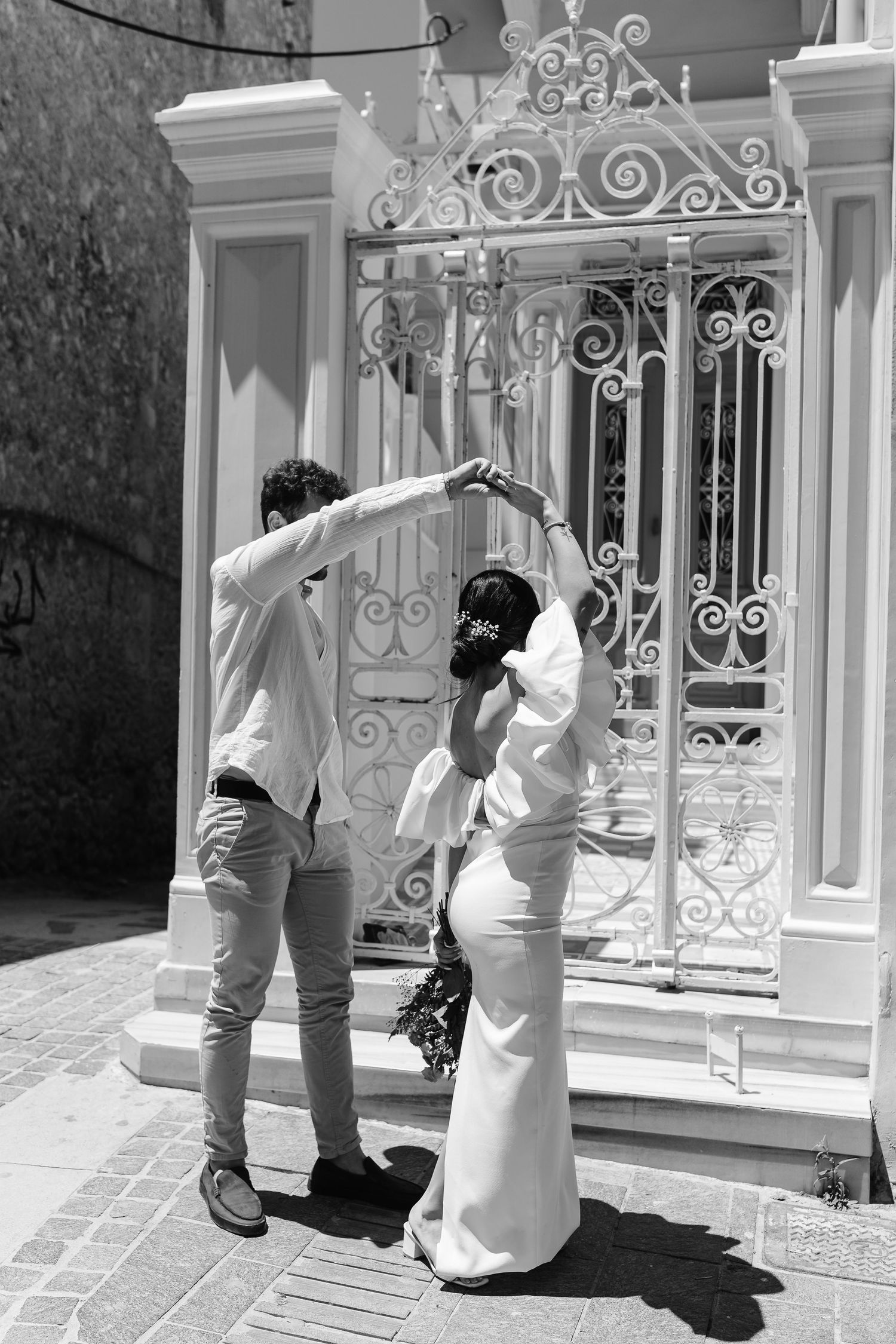 Black and white photo of couple dancing near decorative white iron gate, groom spinning bride in wedding dress.