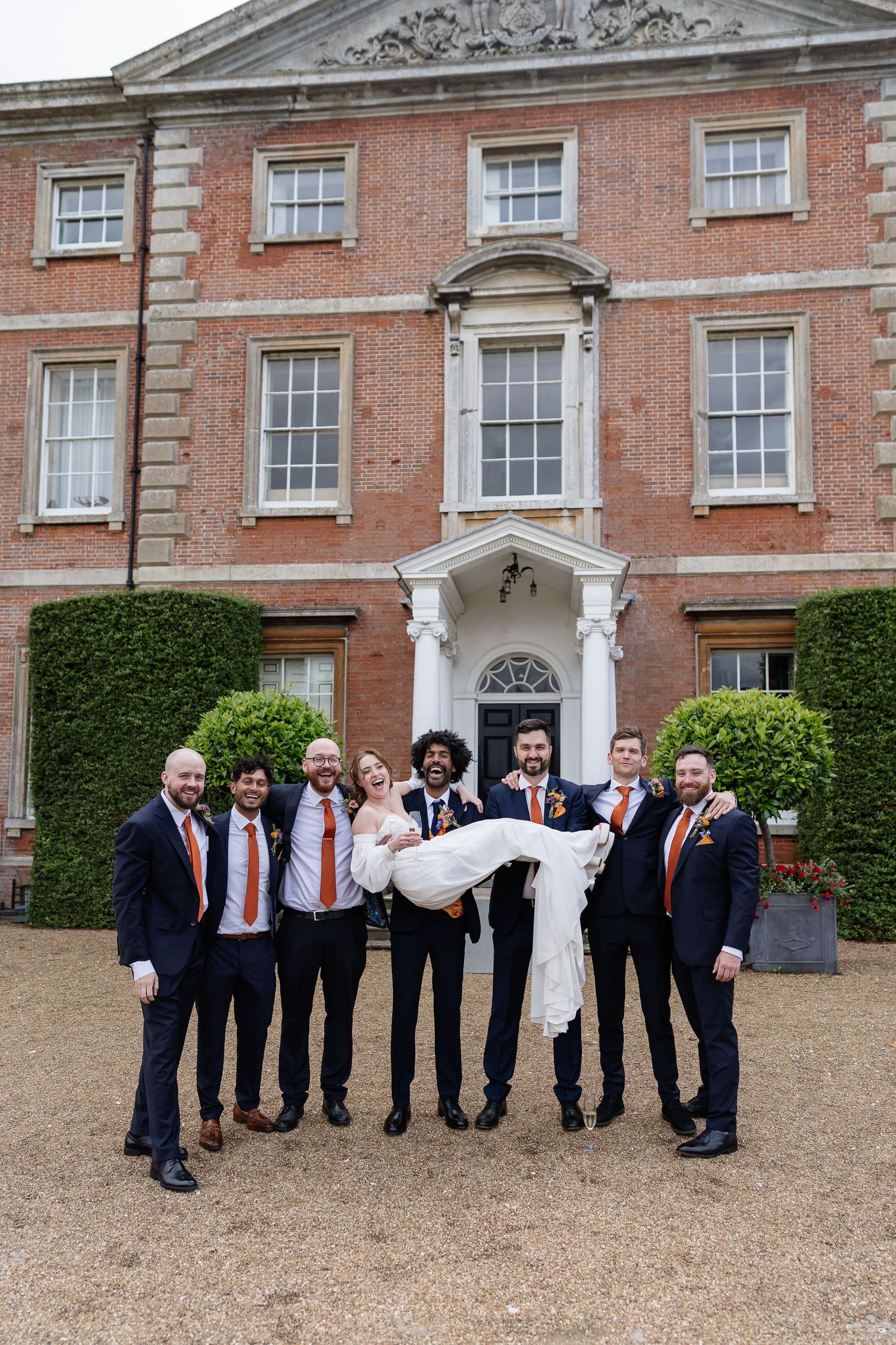 Groom and groomsmen in navy suits with orange ties lift smiling bride outside elegant brick manor.