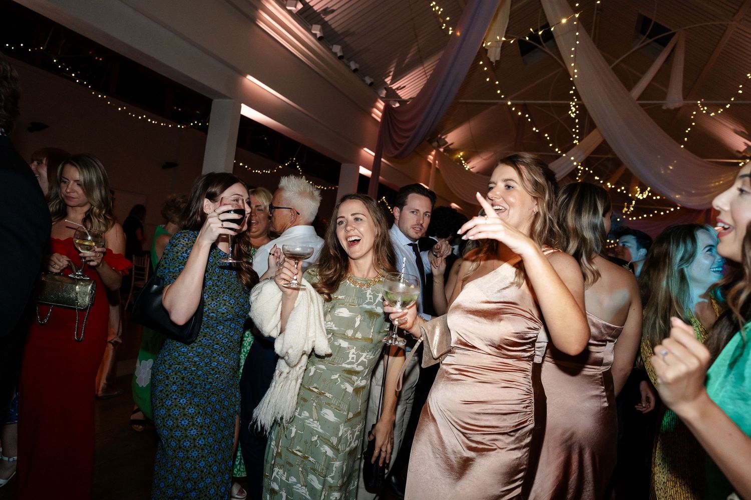 Wedding guests laugh and dance energetically on decorated dance floor with fairy lights at evening reception.