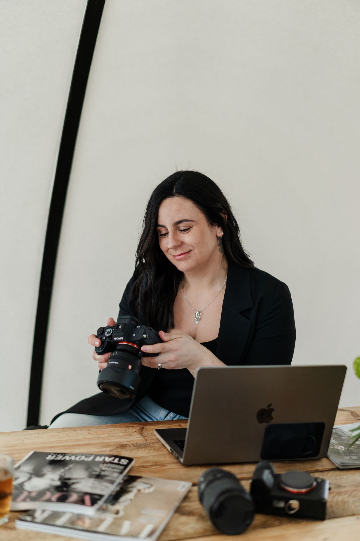 Woman photographer smiling while holding a DSLR camera at a wooden desk with a MacBook and lenses.