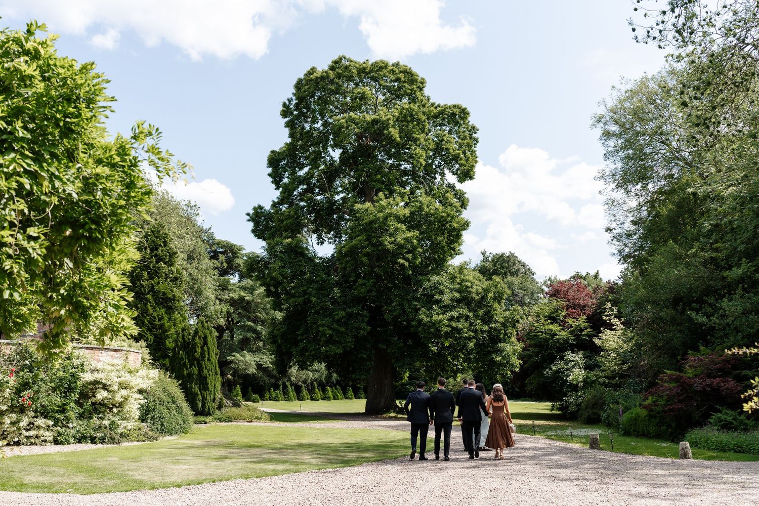 Wedding guests walking along gravel path through lush green English country garden on sunny day.