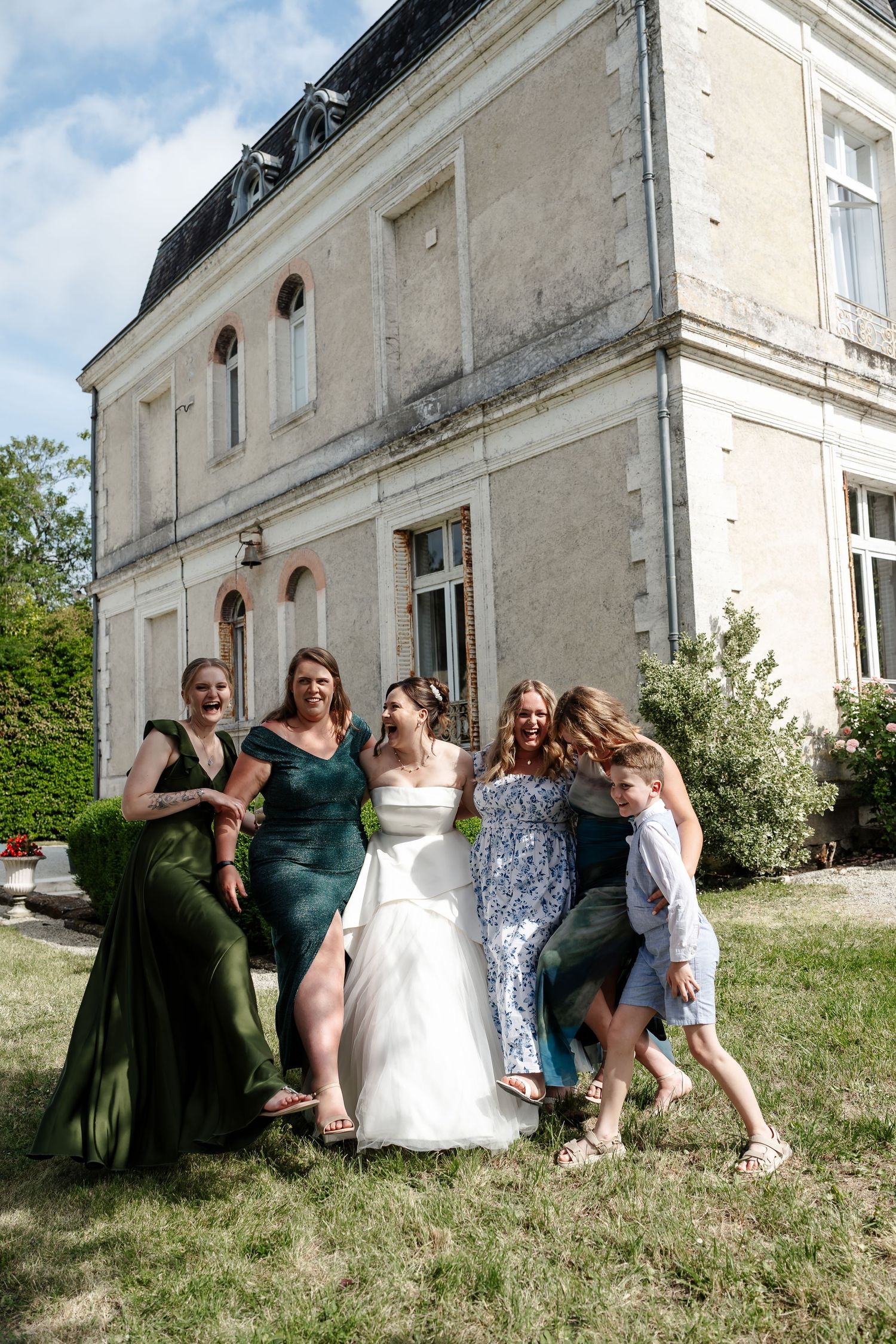 Bride in white gown poses with bridesmaids in green dresses outside a French chateau on a sunny day.