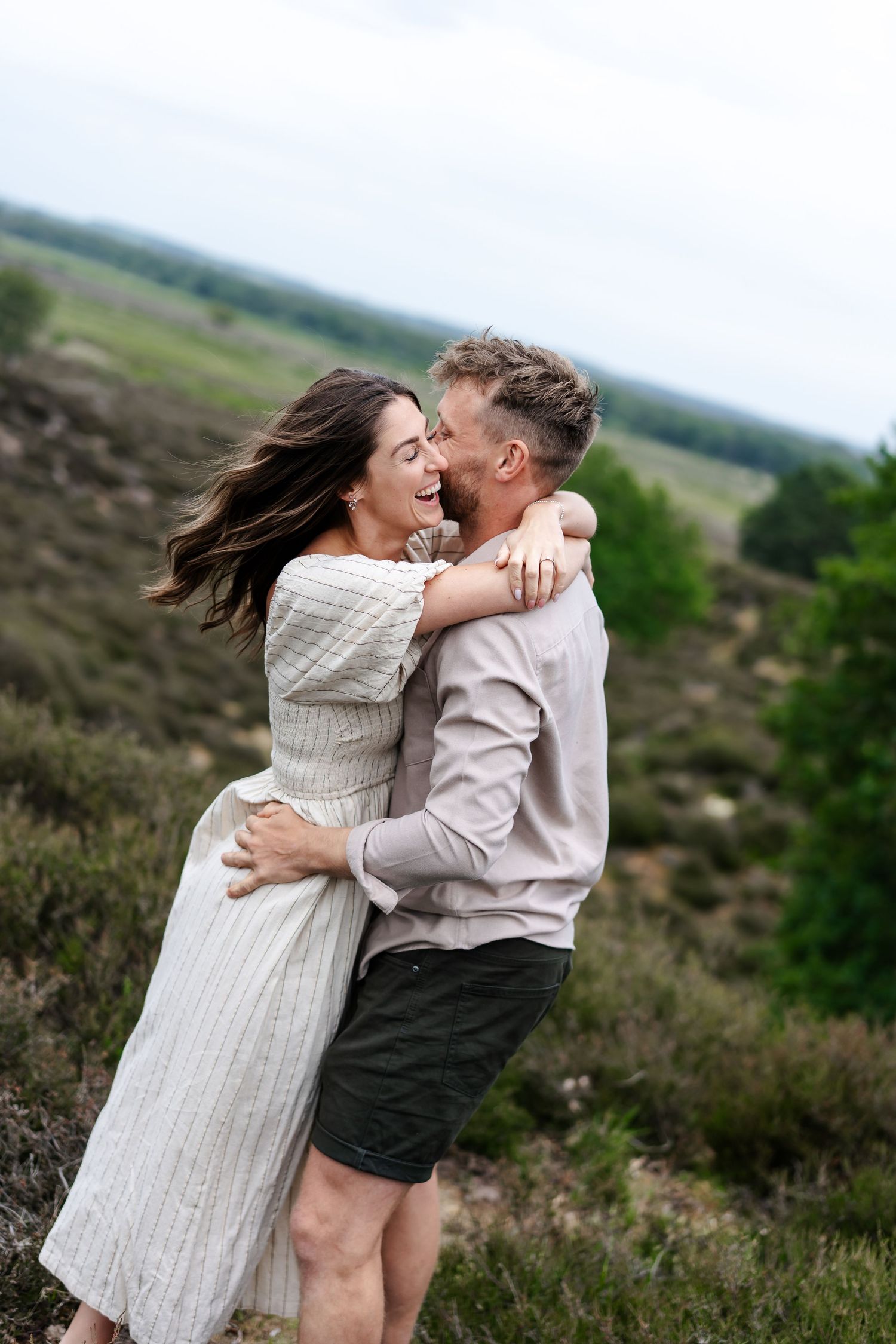 A joyful couple embraces and laughs together on a scenic heathland landscape with rolling hills behind.