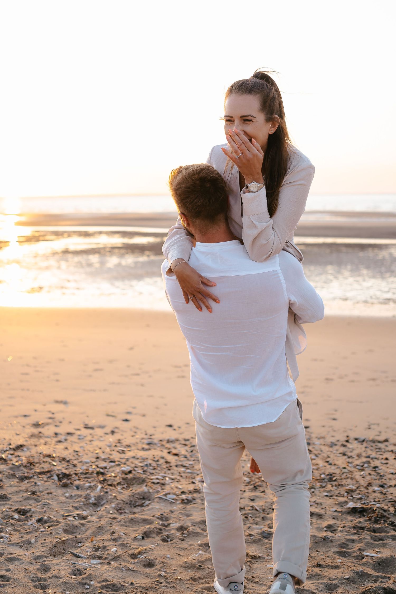 Man lifting woman on beach at sunset as she covers her mouth in surprised joy.