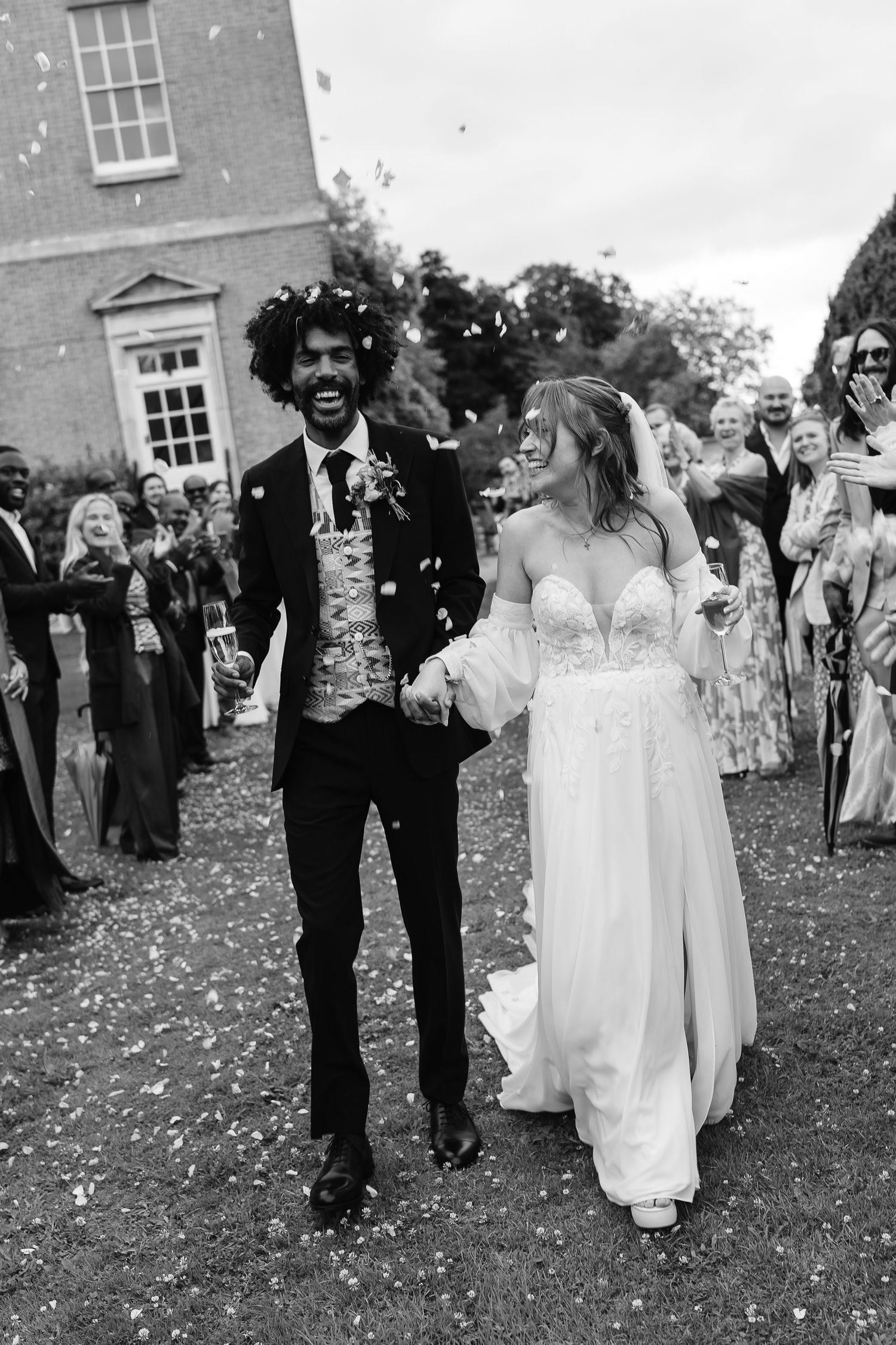 Black and white photo of laughing bride and groom walking through confetti with wedding guests.