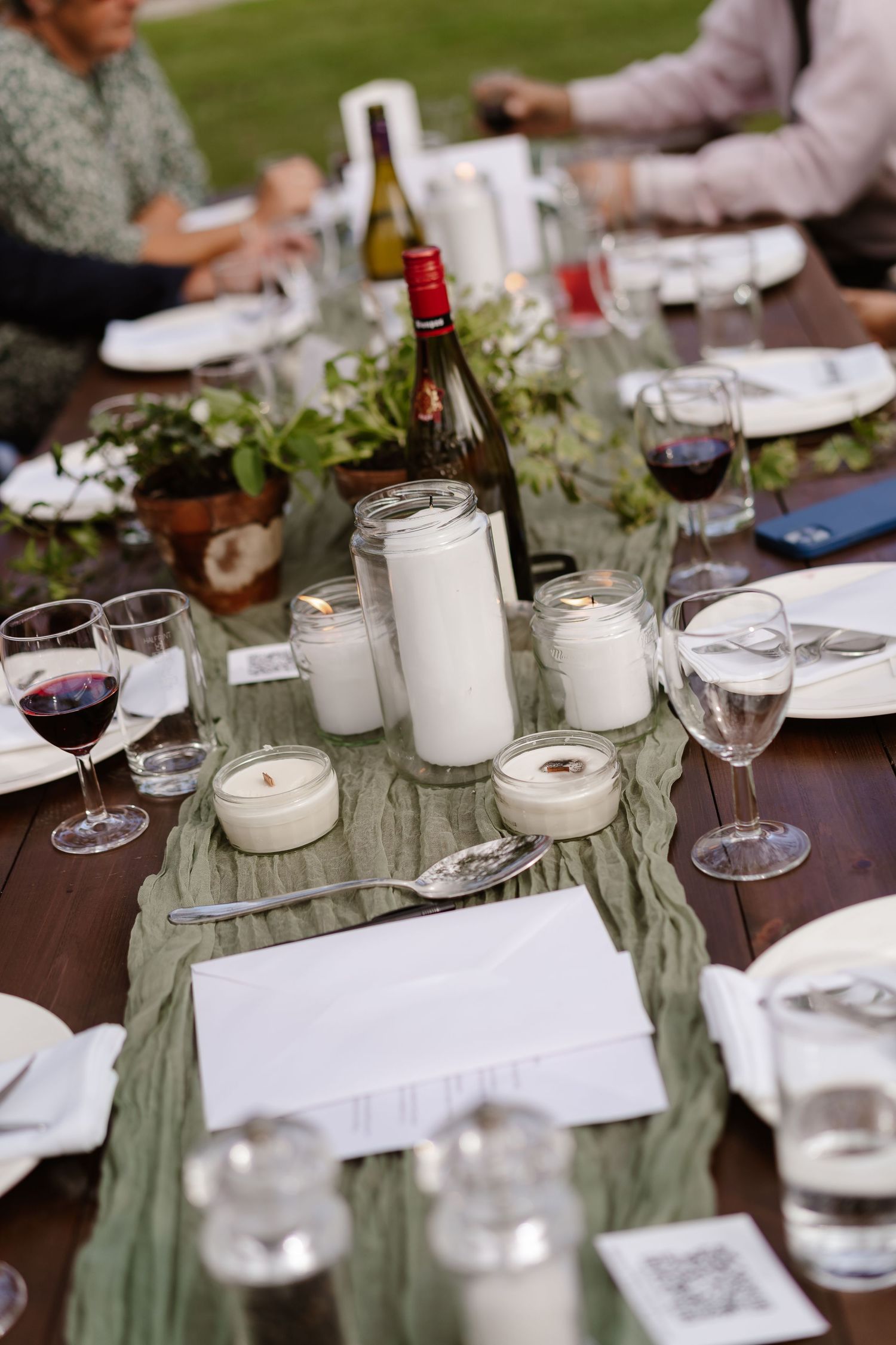 Elegant outdoor wedding reception table with candles, greenery runner, wine glasses, and red wine bottle on rustic wood table.