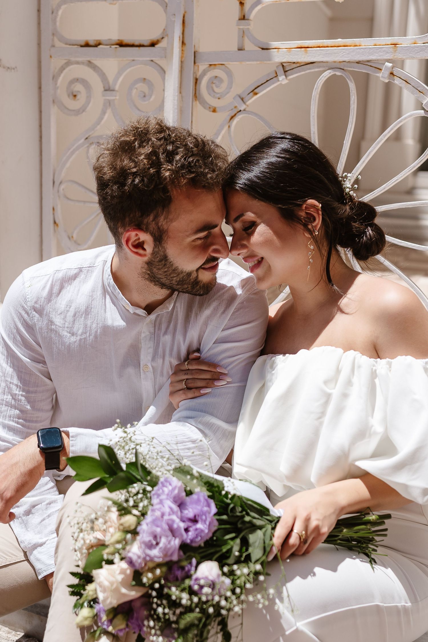 Bride and groom sitting close together smiling with a purple and white wedding bouquet in sunlight.