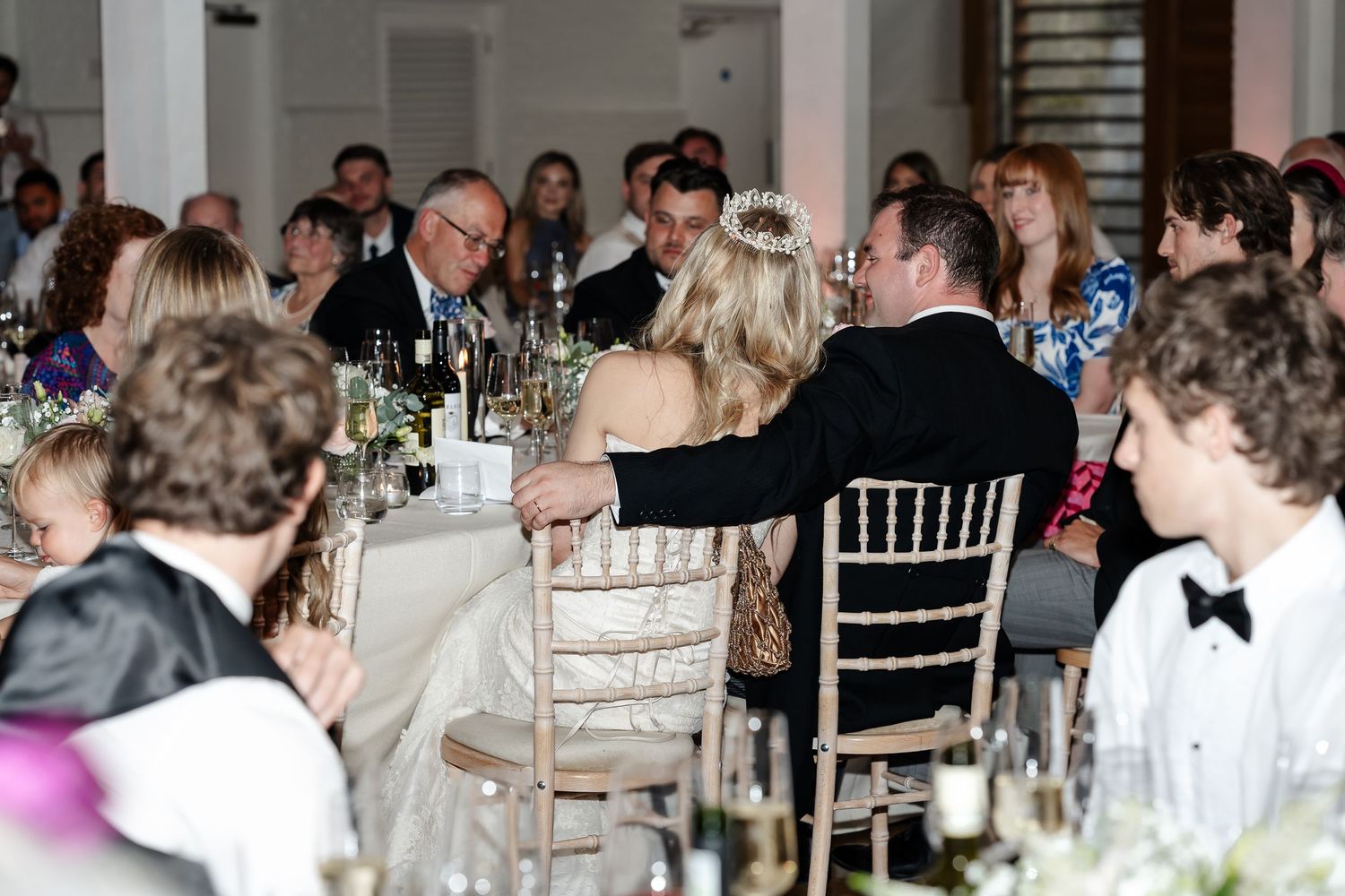 Elegant wedding reception dinner with guests seated at long tables in a formal venue, bride and groom sharing a romantic moment.