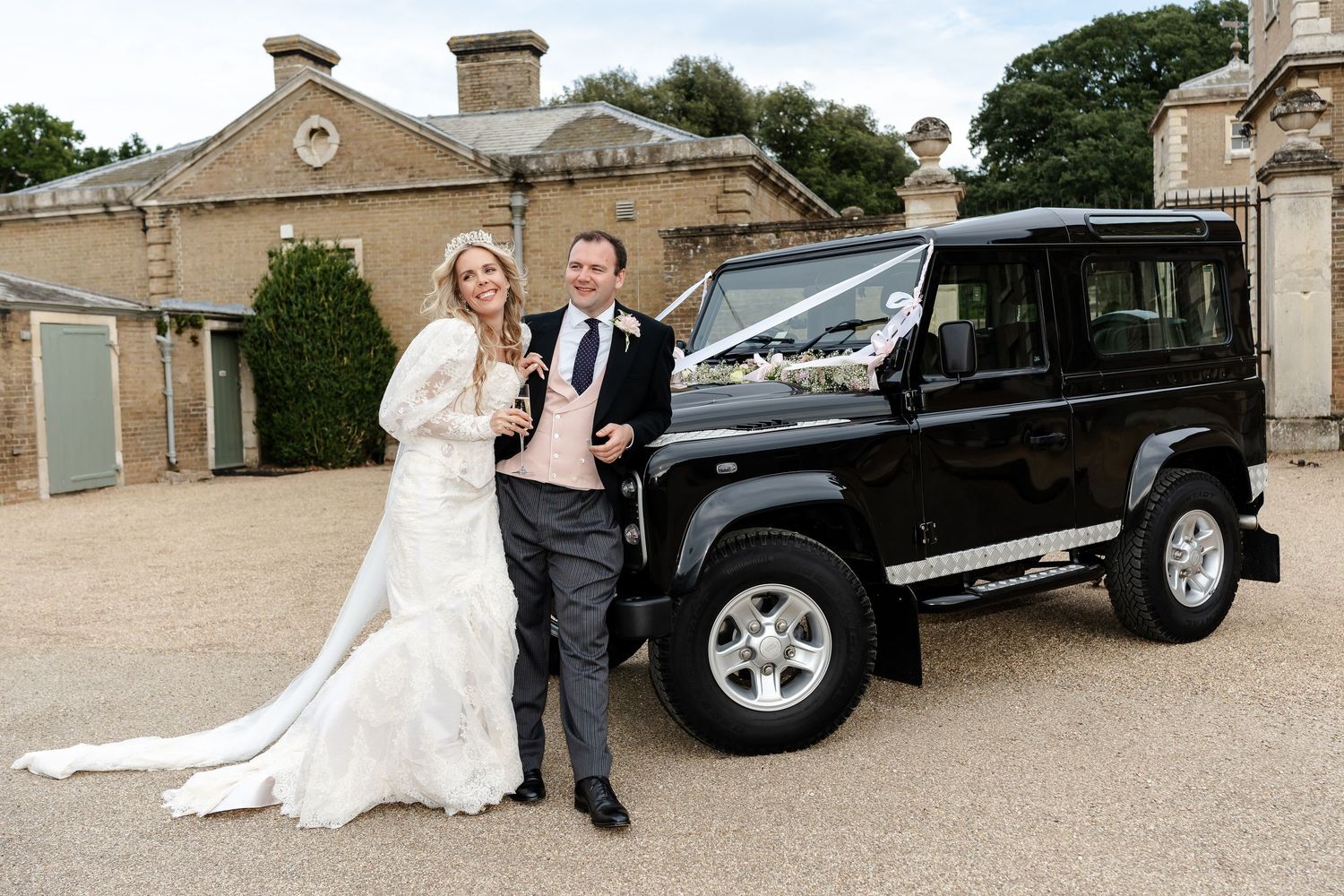 Bride and groom posing with a black Land Rover Defender decorated with white ribbons outside a stone building.