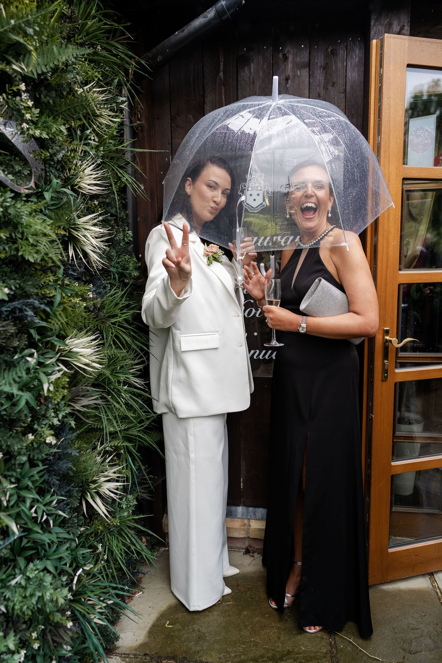 Bridesmaid in white suit and guest in black dress shelter under clear umbrella at wedding.