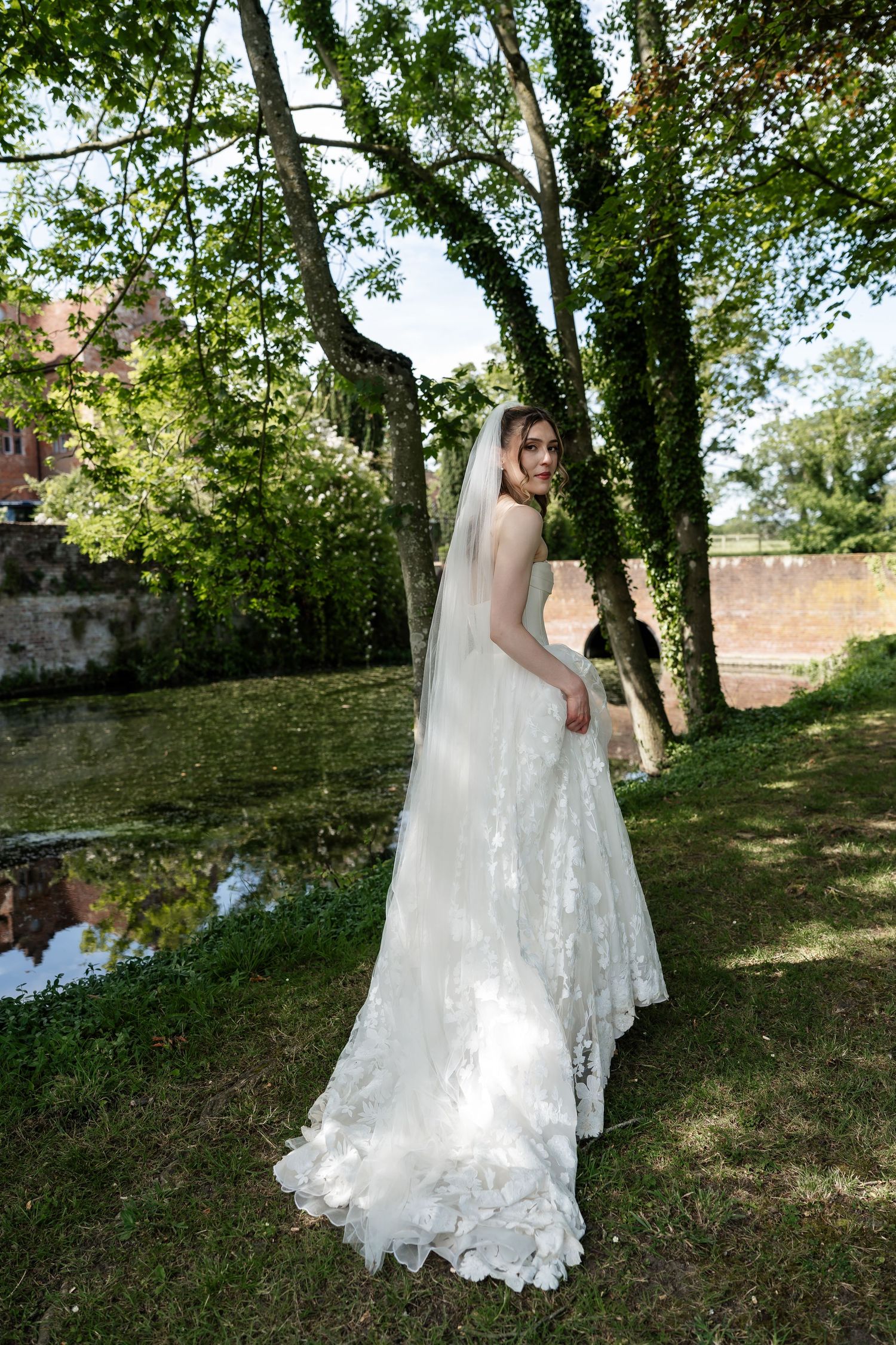 Bride in lace gown and veil stands among trees beside a pond at historic venue.