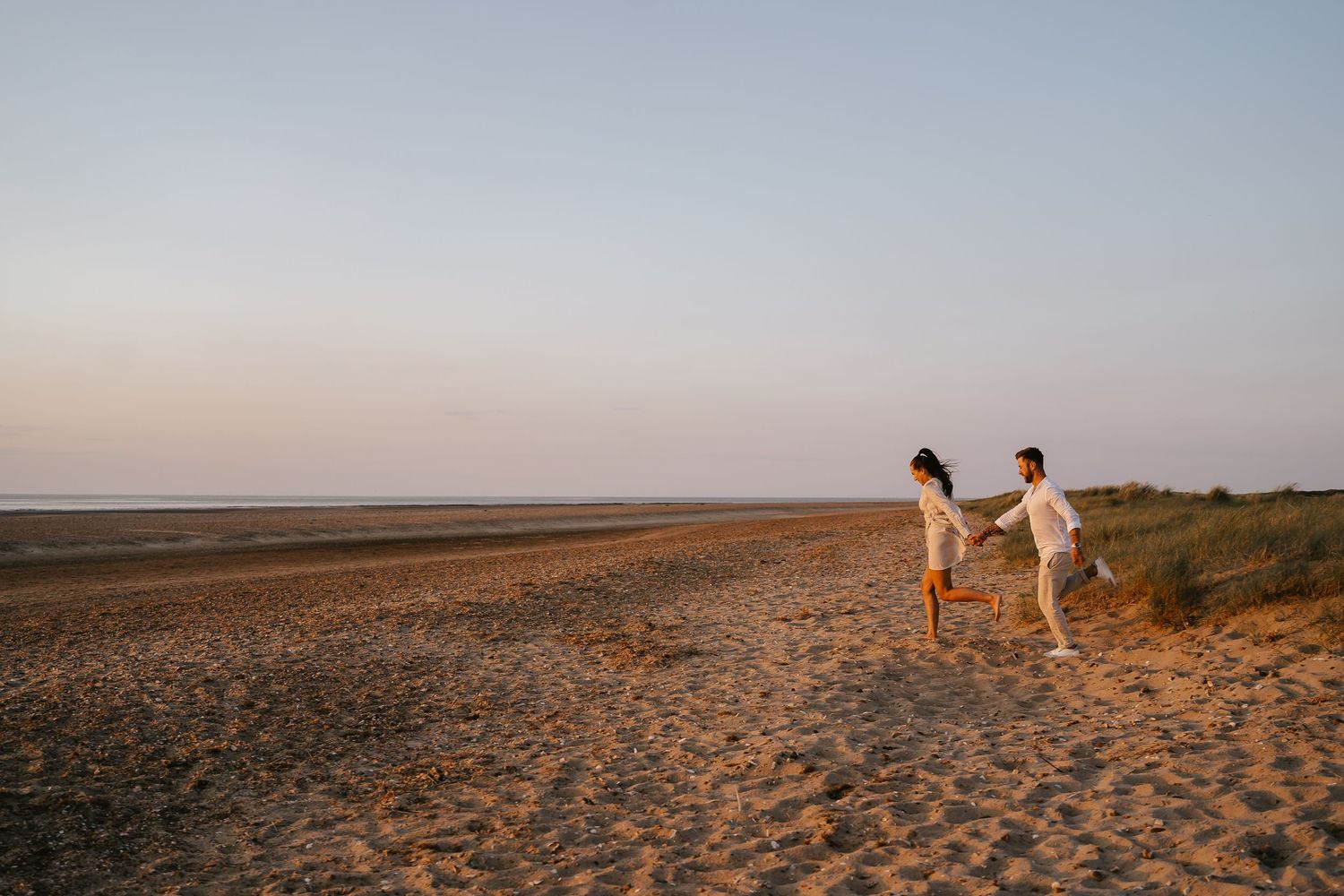 Couple dressed in white playfully running hand in hand along a wide sandy beach at sunset.