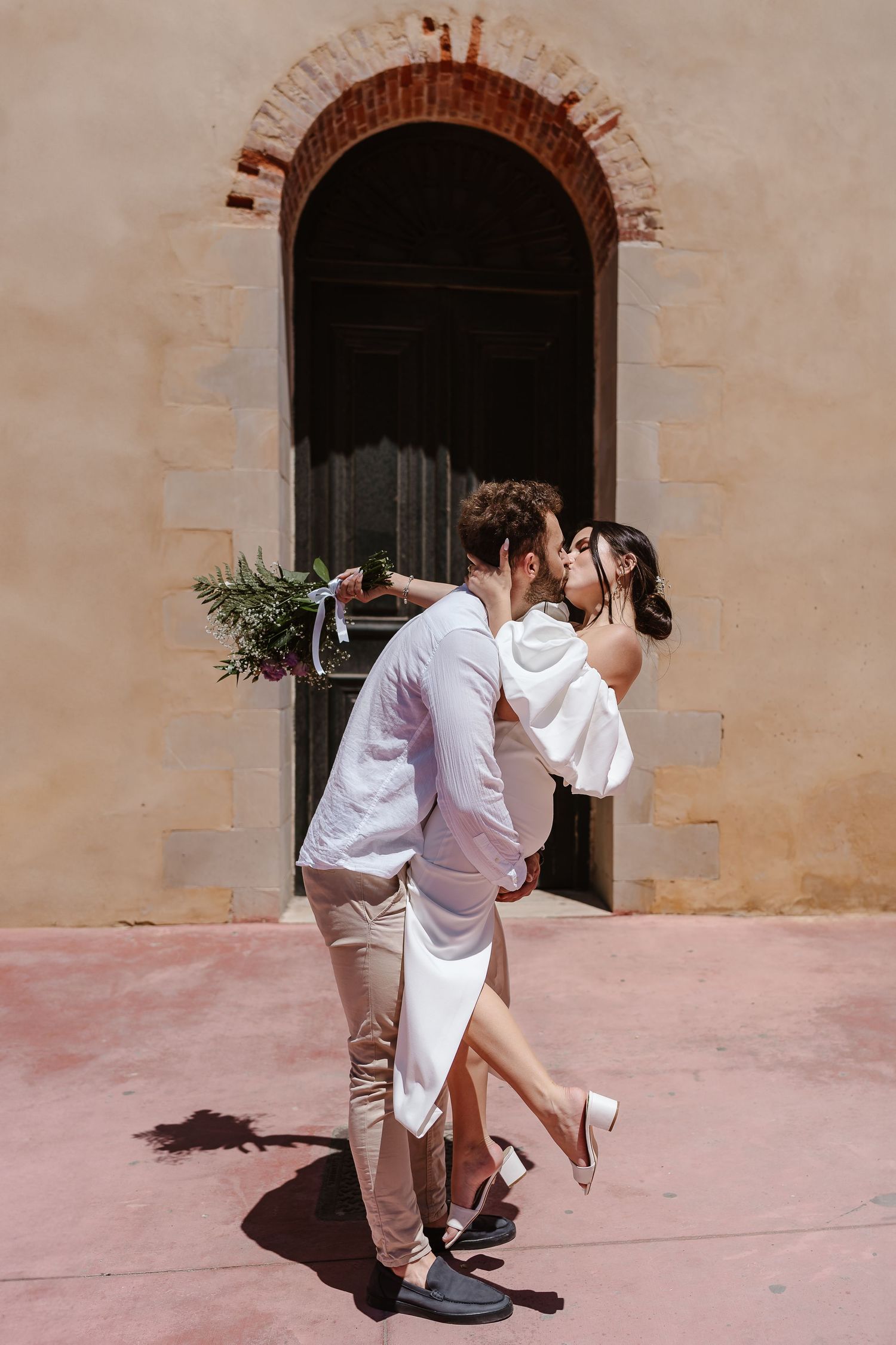 Bride and groom share a romantic kiss outside a rustic arched doorway, bride holding a floral bouquet.