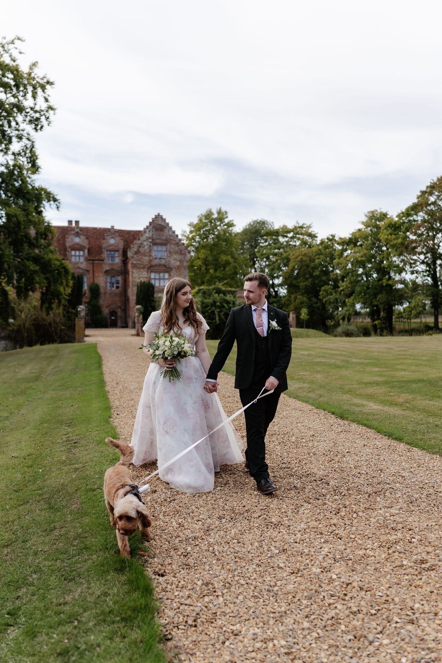 Smiling bride and groom walk their dog along a gravel path outside a grand manor house.