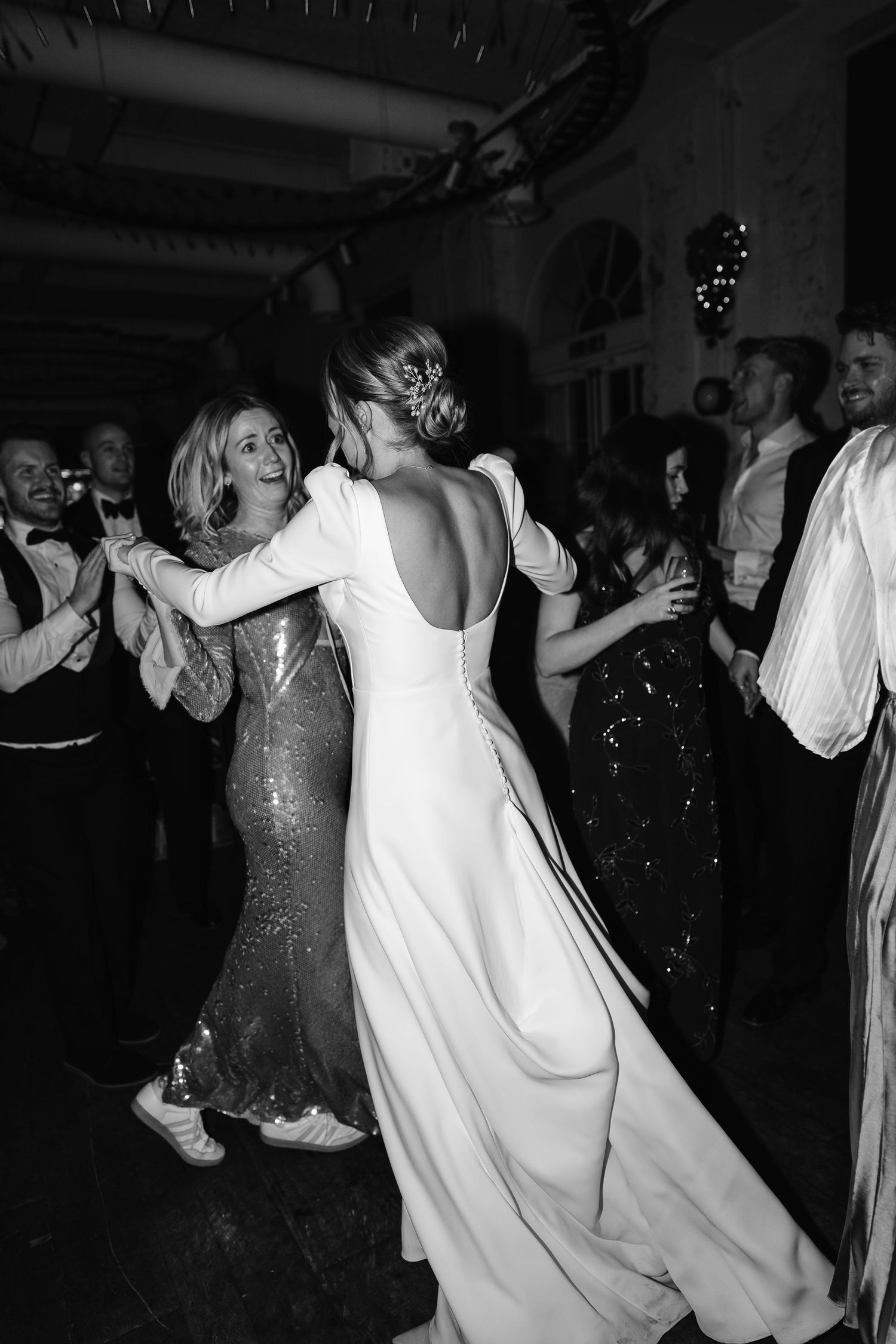Black and white photo of bride in backless white gown dancing with guests at wedding reception.